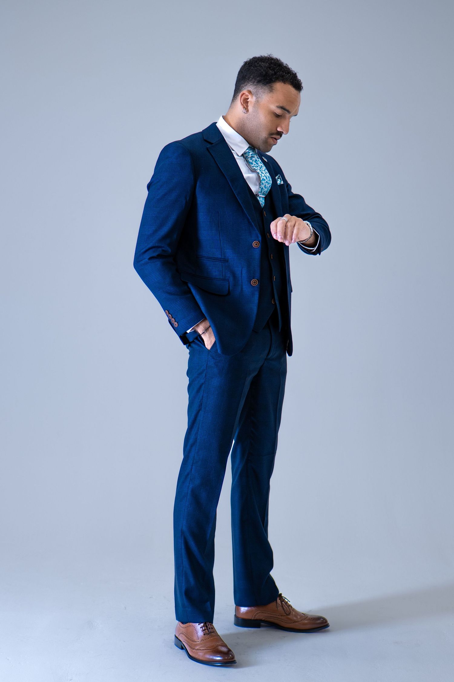 Man in navy three-piece suit checking his watch, wearing brown leather Oxford shoes.