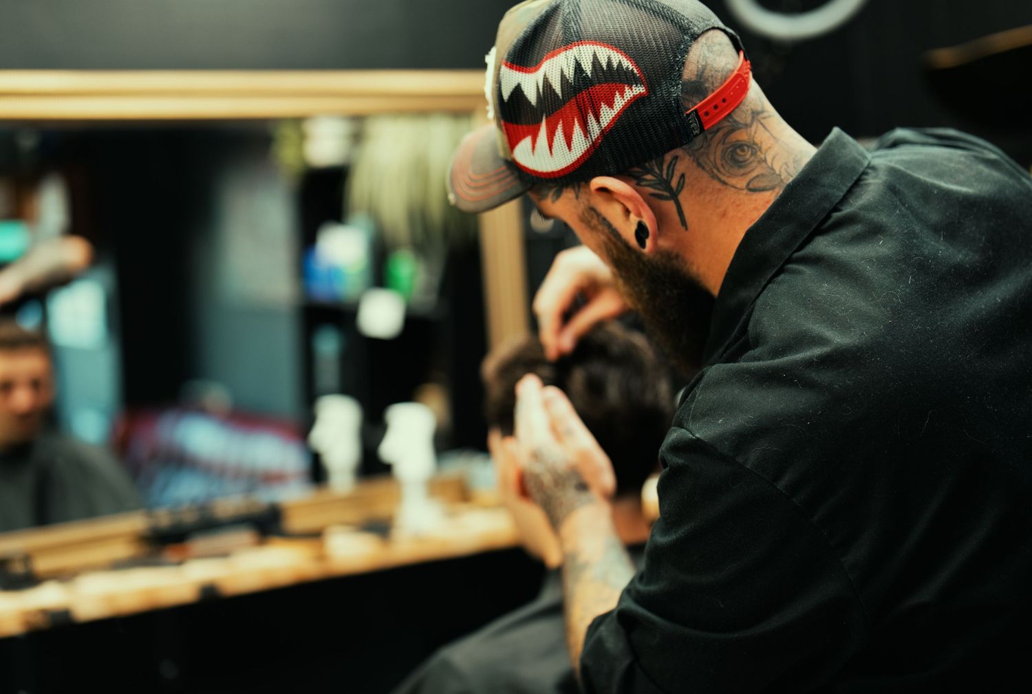 Close-up of a tattooed barber wearing a shark-print cap focusing on a client's haircut in a barbershop.