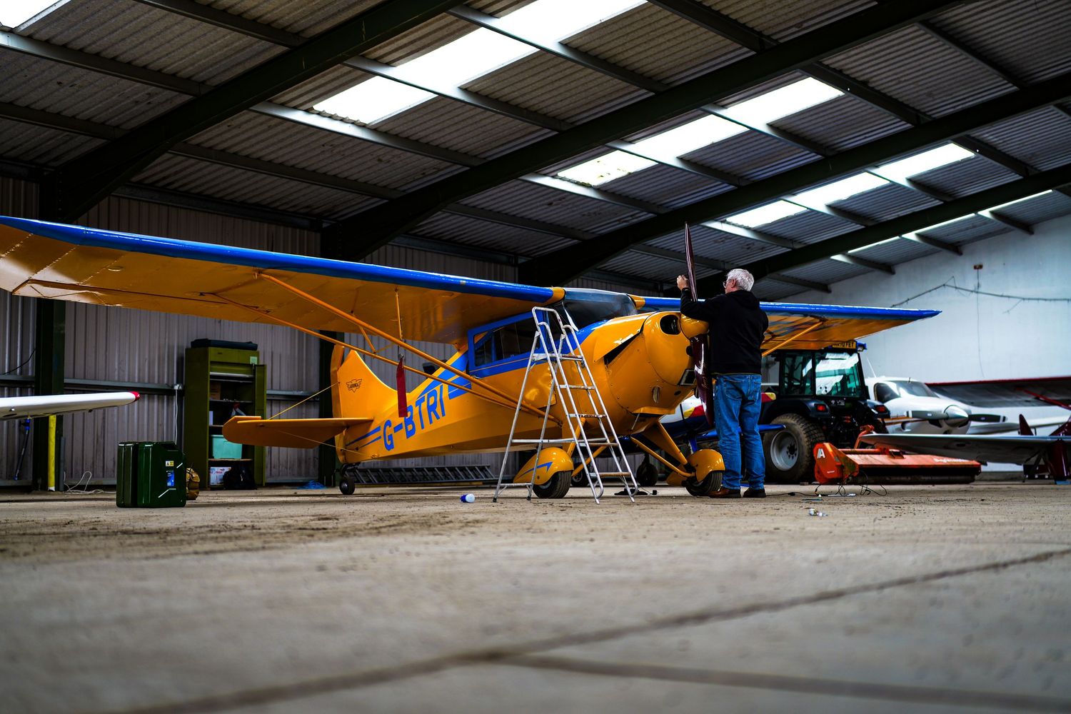 Yellow and blue light aircraft undergoing maintenance in a spacious hangar with a ladder nearby.