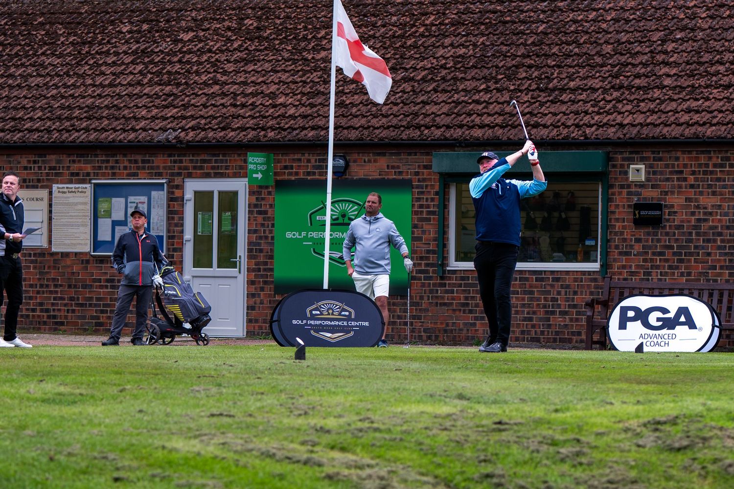 Golfer teeing off near a brick clubhouse with PGA and England flag visible.