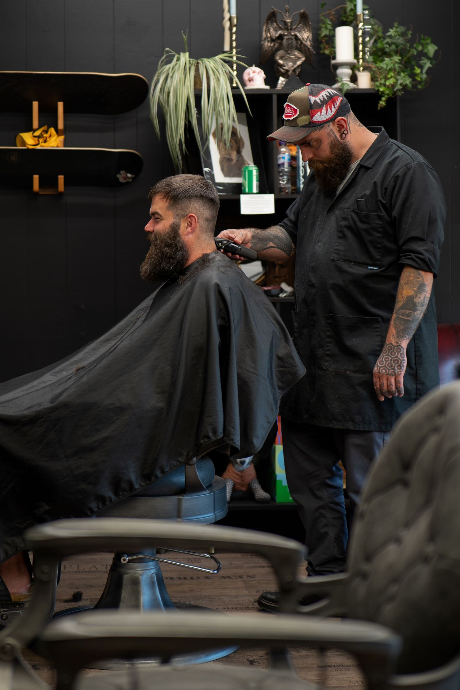 Tattooed barber in a cap trimming a bearded client's hair in a stylish dark barbershop.