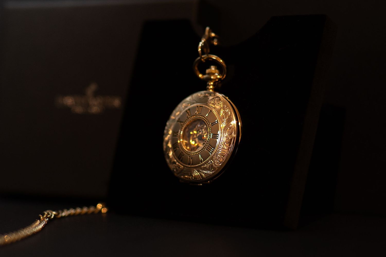 Ornate gold pocket watch on display stand with decorative engraved case and chain against dark background.