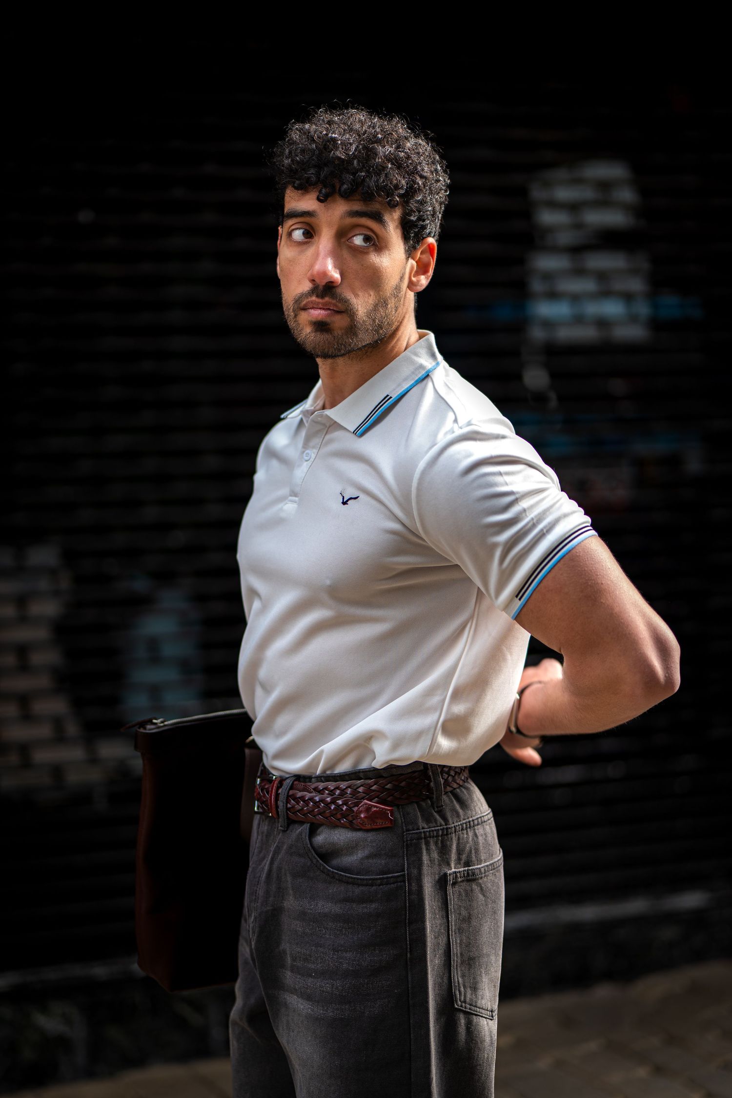 Curly-haired man in white polo shirt holding folder posed against dark brick wall background.