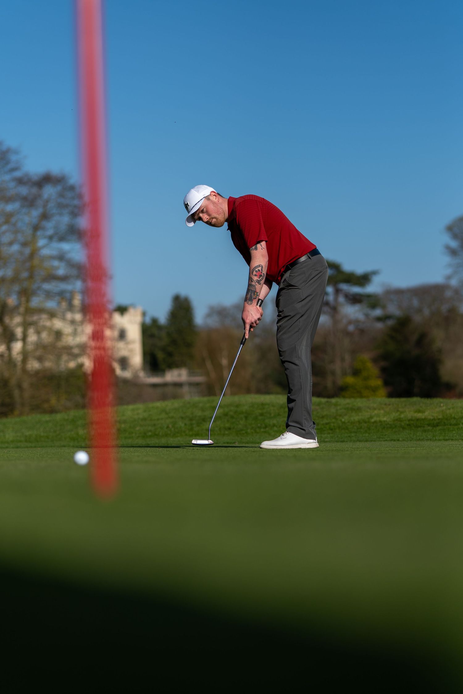 Tattooed golfer in red shirt putting on green with red flag in foreground.