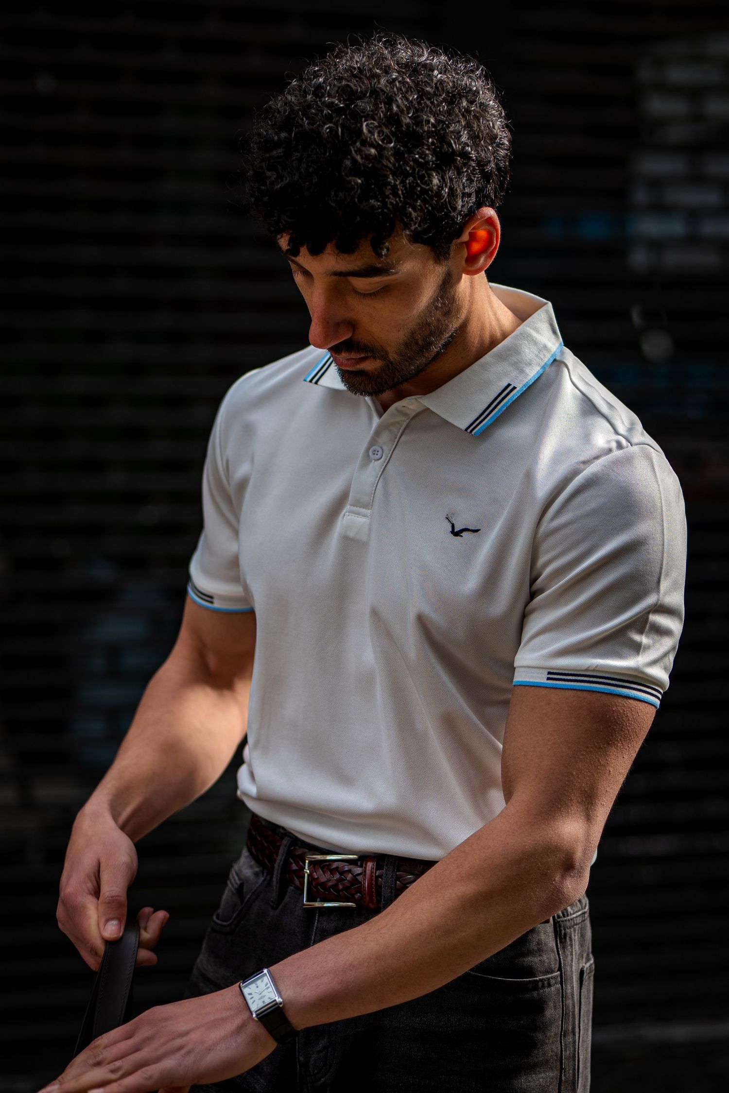 Muscular man in white polo shirt with blue trim and bird logo looking down against dark background.