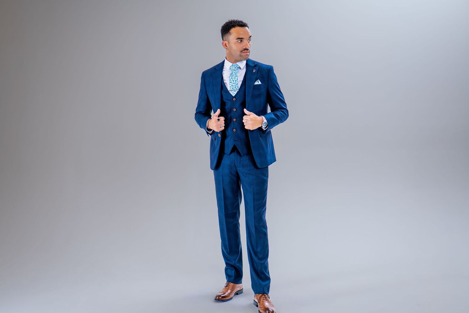 A man poses confidently in a tailored navy blue three-piece suit with a light blue tie on grey background.