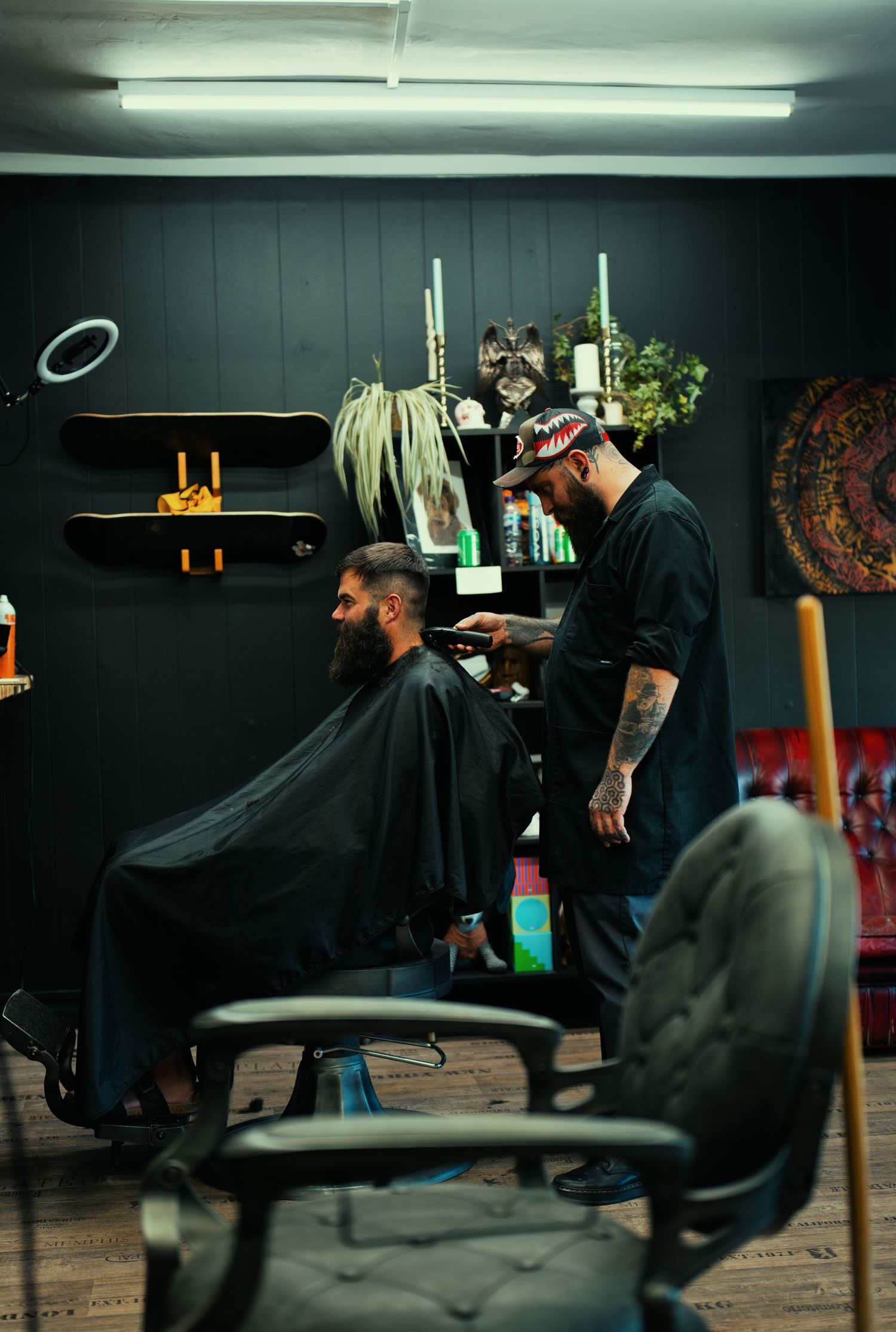 Tattooed barber cutting bearded mans hair in stylish dark barbershop with shelves of products.