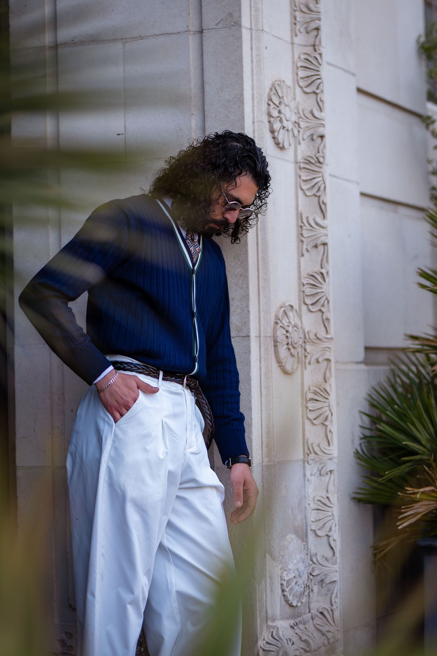 Stylish man in navy pinstripe cardigan and wide-leg white trousers leaning against decorative stone wall.