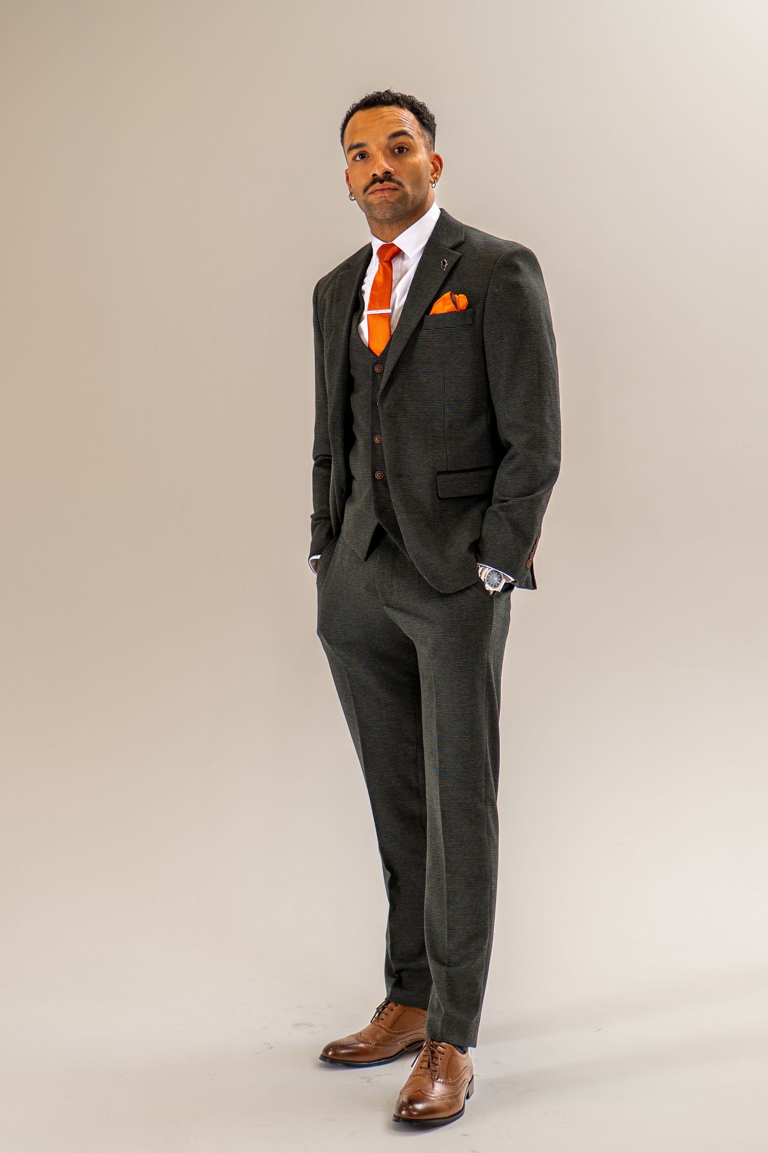 Stylish man in a dark three-piece suit with orange accessories and brown brogues standing tall.