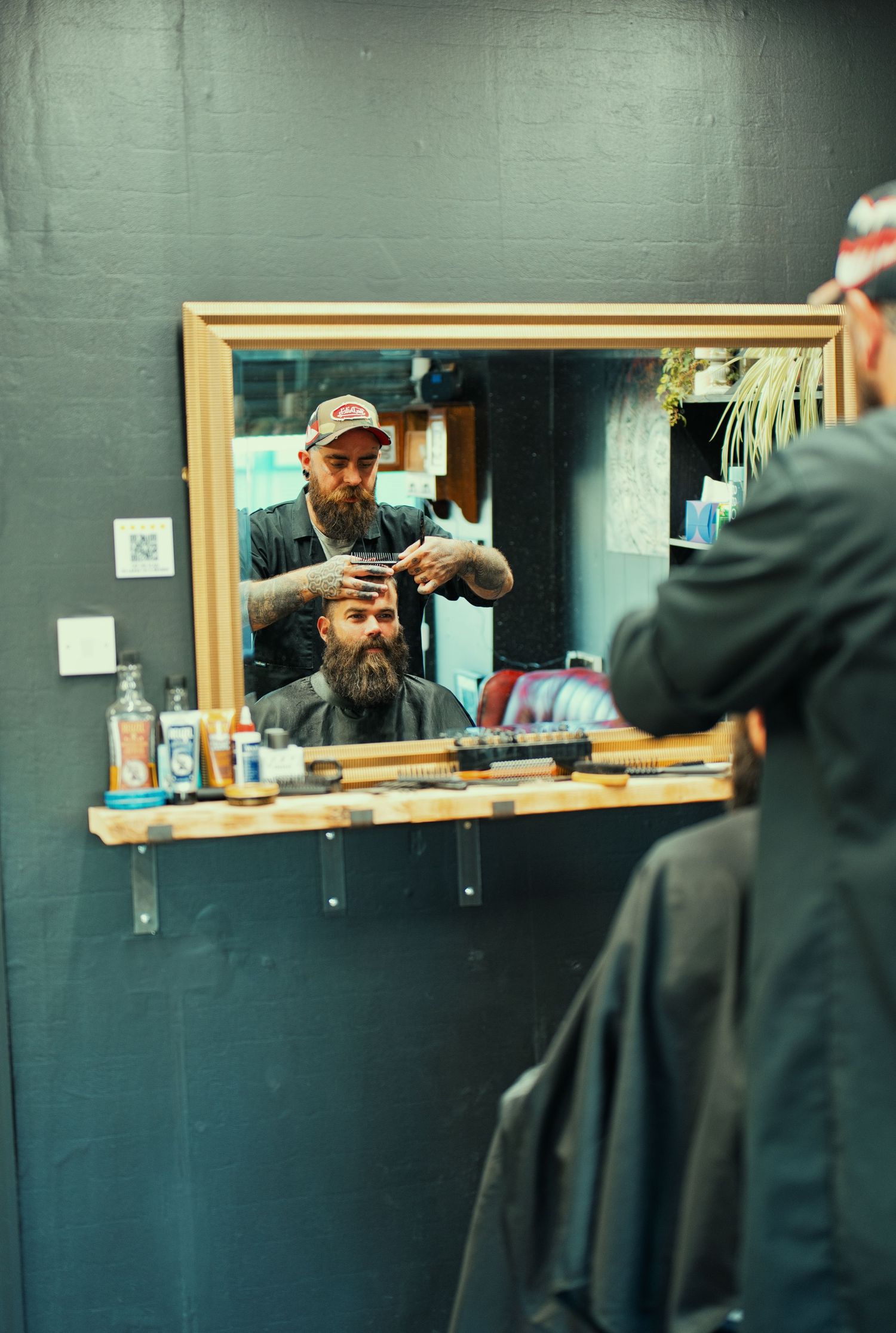 Tattooed barber cutting a bearded man's hair in a barbershop, reflected in a gold-framed mirror.