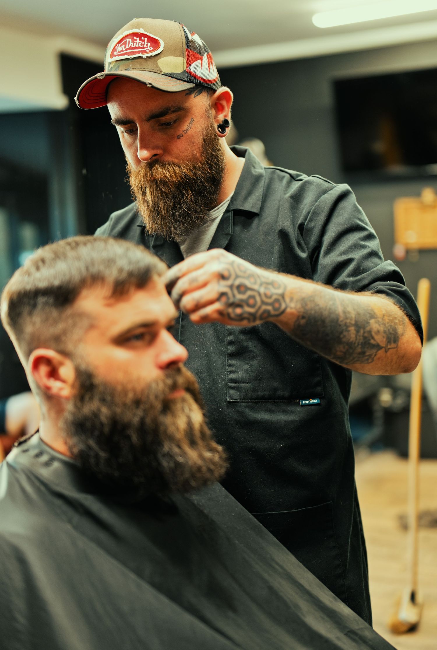 A bearded tattooed barber wearing a cap carefully cuts a bearded client's hair in a barbershop.