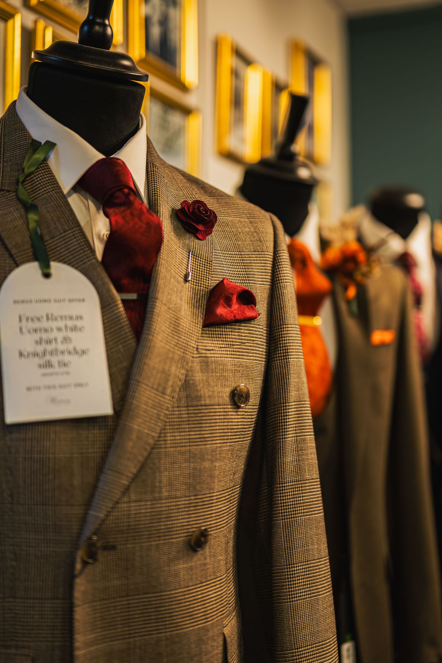Row of tailored tweed suits on mannequins with red rose accessories and pocket squares in an upscale menswear shop.