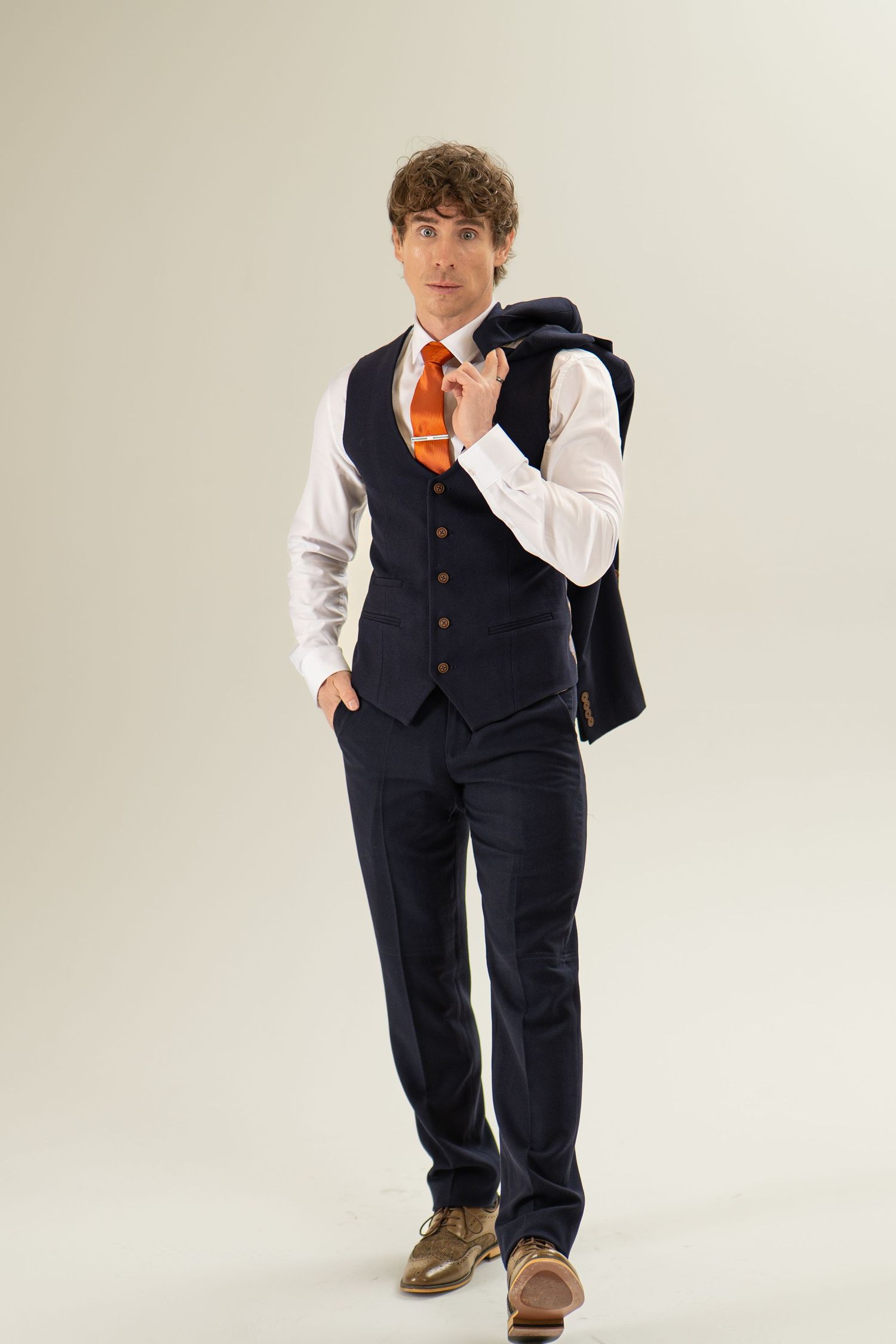 Stylish man in navy suit vest and orange tie carrying jacket over shoulder, walking.