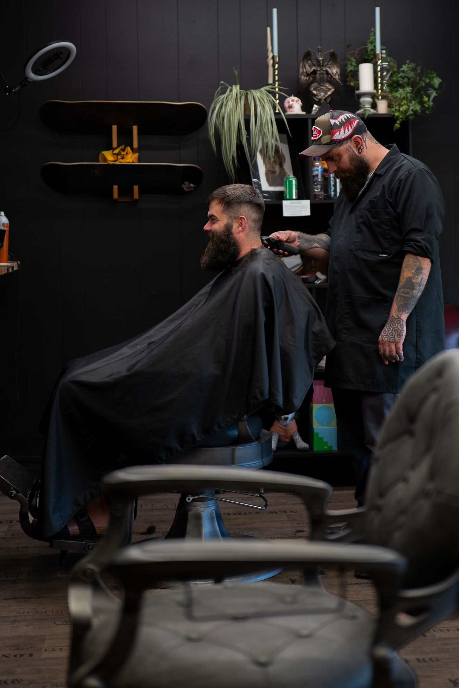 Tattooed barber cutting bearded man's hair in a modern dark-themed barbershop.