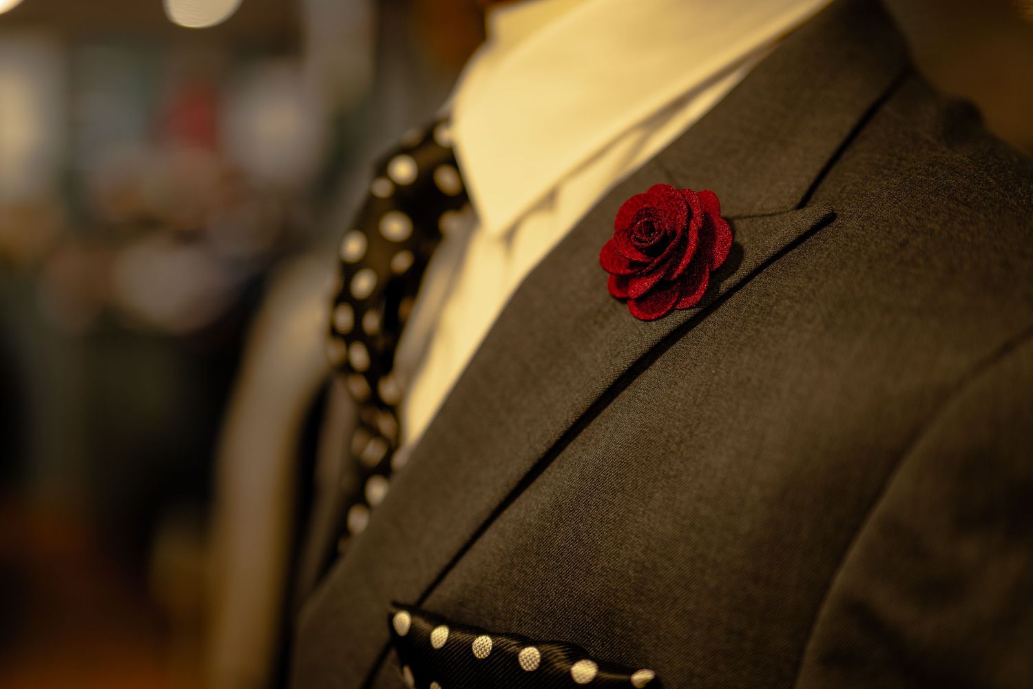 Close-up of a dark suit jacket on a mannequin featuring a red rose lapel pin and polka dot tie.