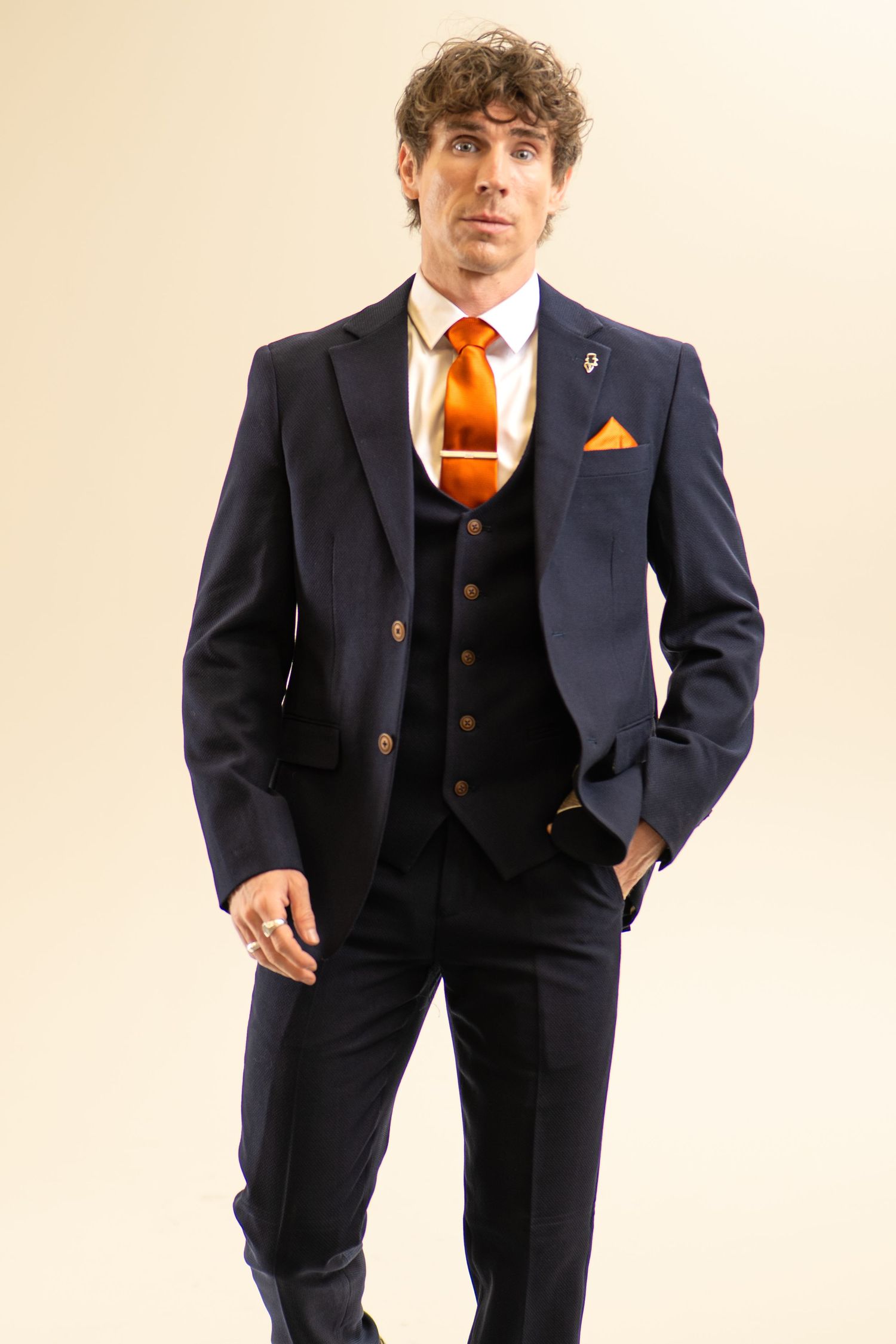 Man posing in navy blue three-piece suit with vibrant orange tie and gold pocket square in studio.