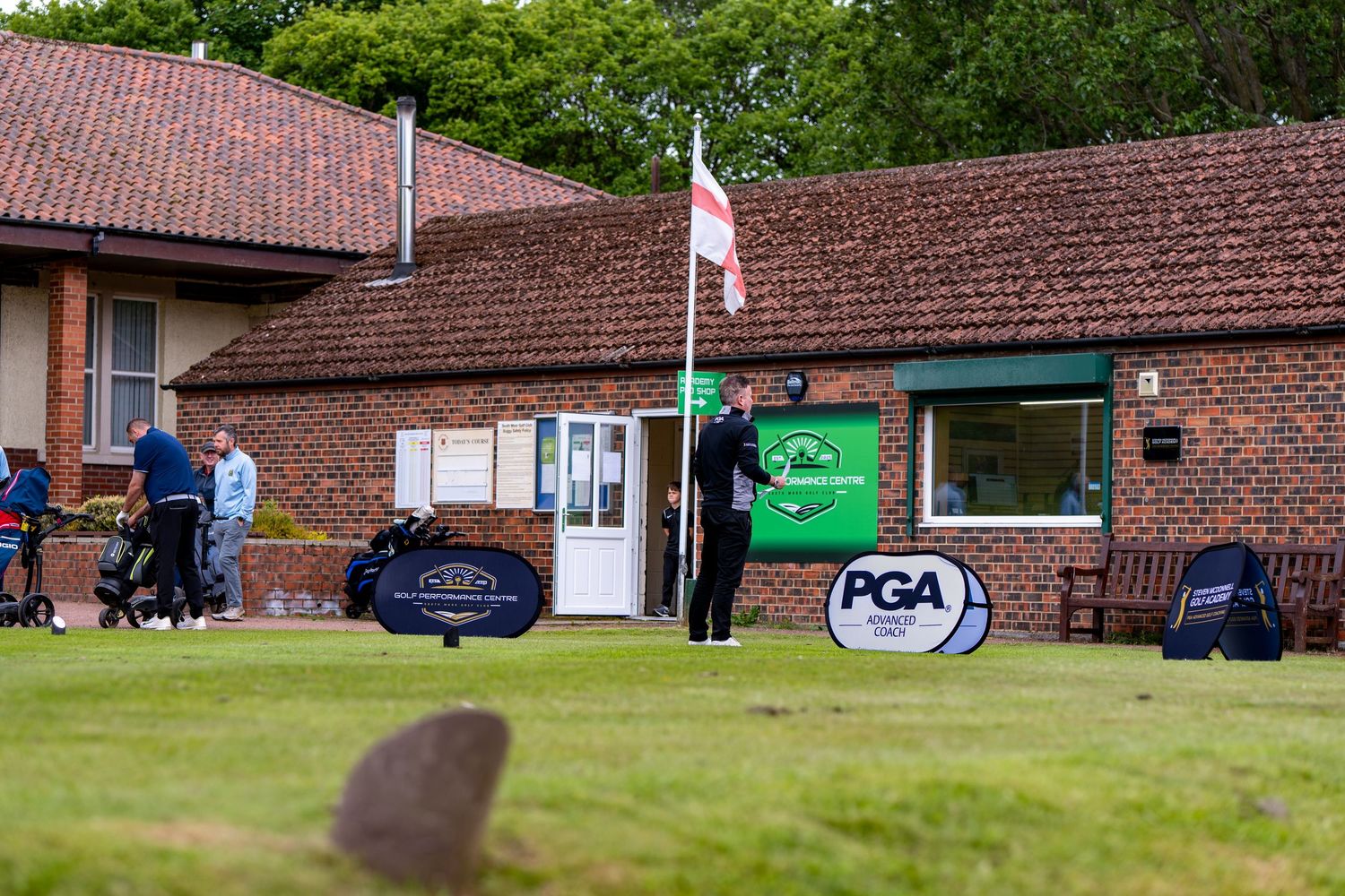 Golfers near a brick clubhouse with PGA banners on a green golf course.