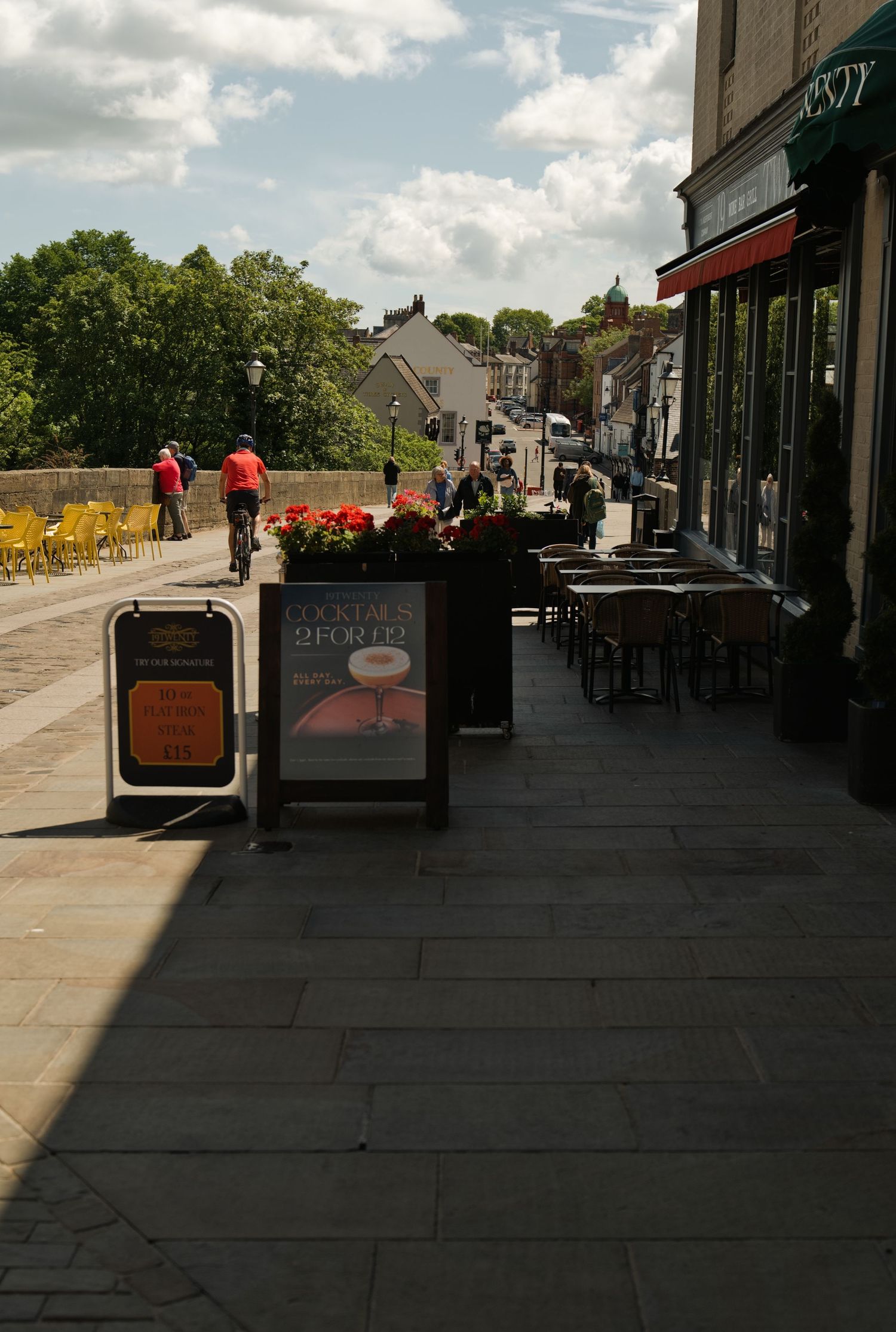 Sunny sidewalk cafe with outdoor seating, cocktail signs, and cyclists on a charming street.