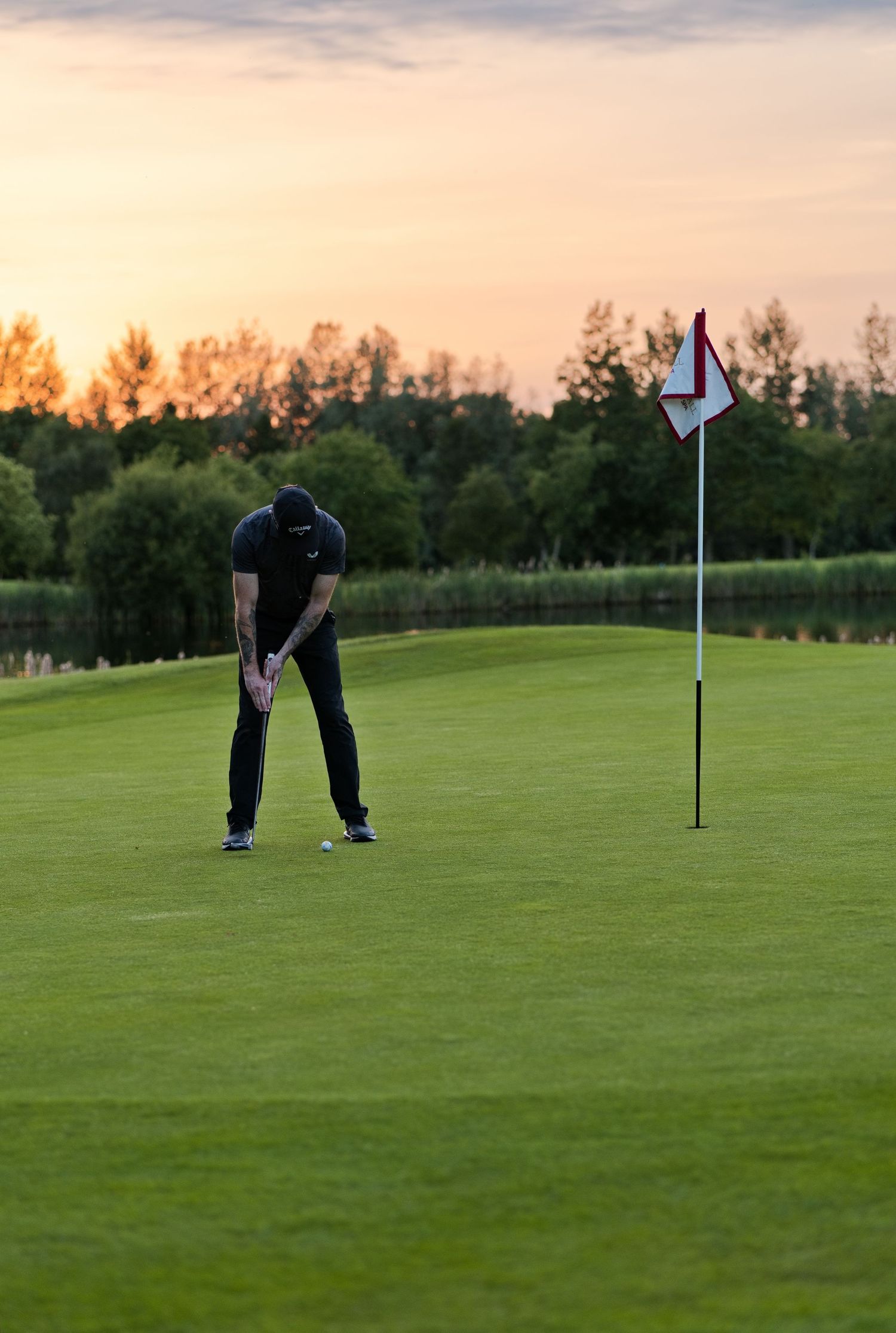 A golfer in all black putts on a golf course green during a stunning golden hour sunset.