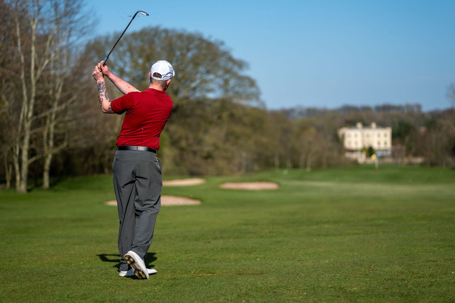 Male golfer finishing a powerful golf swing on a sunny green fairway with trees.