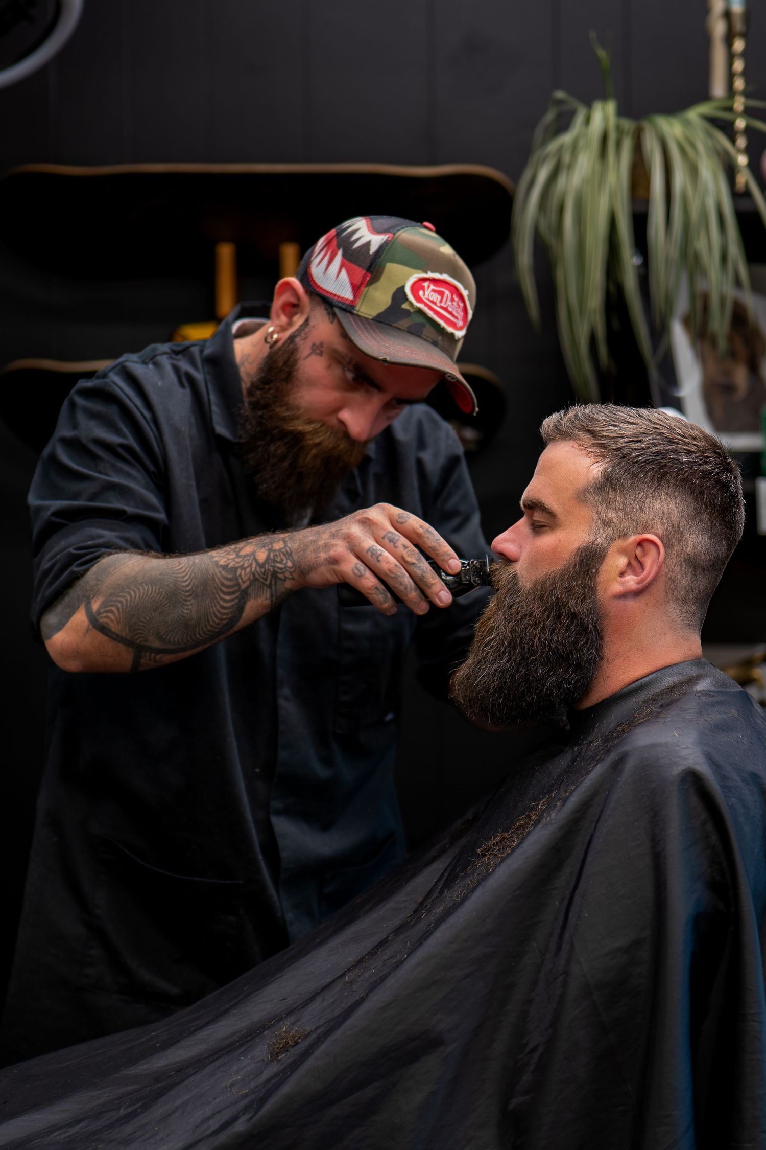 Bearded barber in camo cap trimming client's beard with clippers in dark stylish barbershop.
