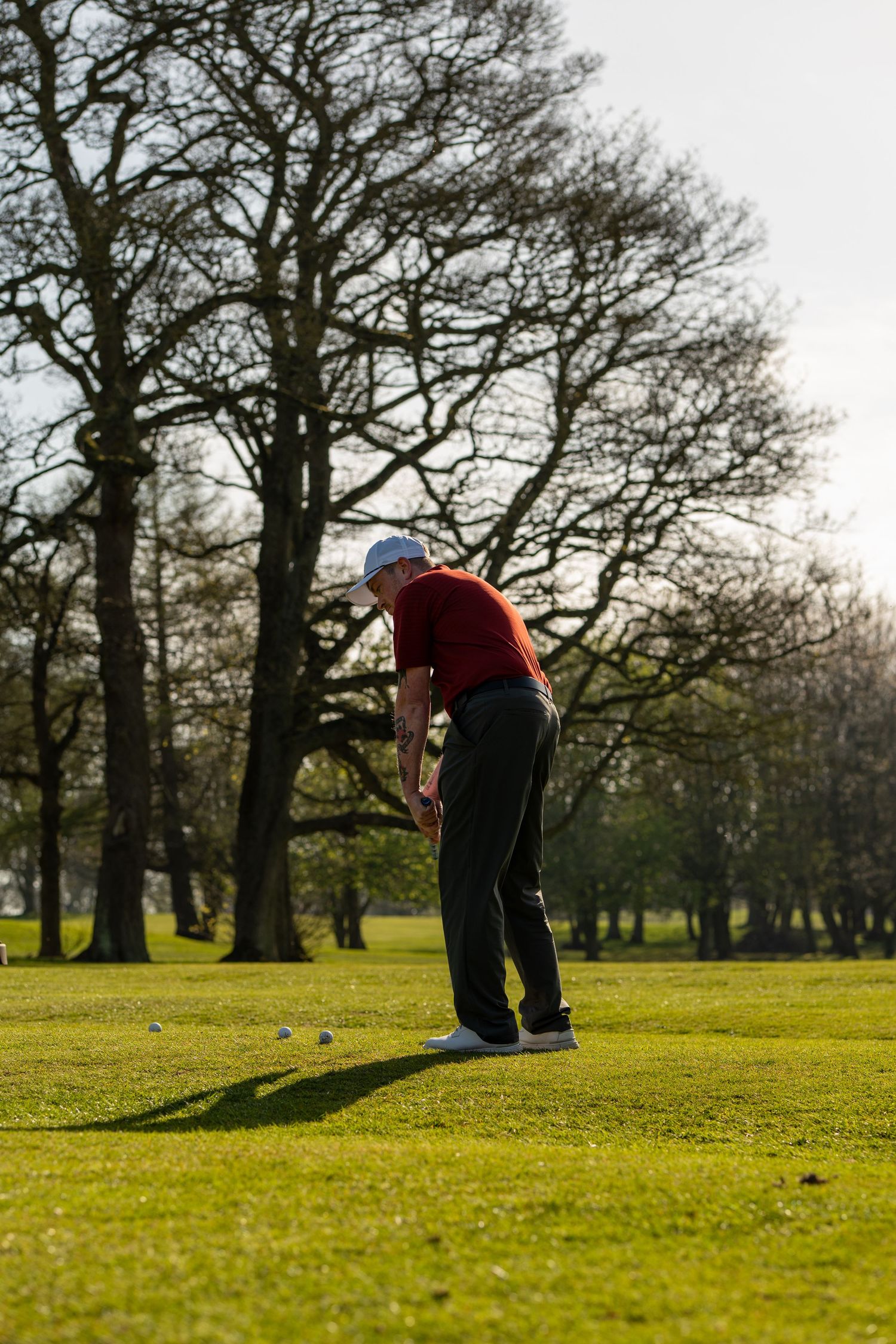 Male golfer in red shirt addressing golf ball on practice area with multiple balls and bare trees in background.