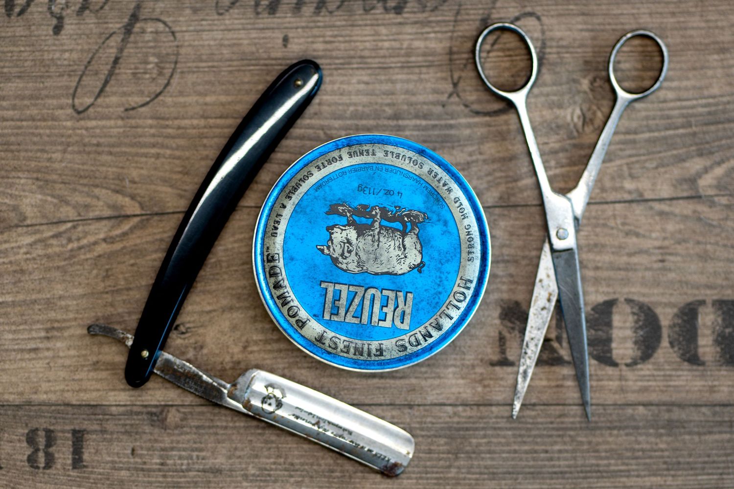 Reuzel blue pomade tin with straight razor and scissors on wooden surface for barber grooming tools.