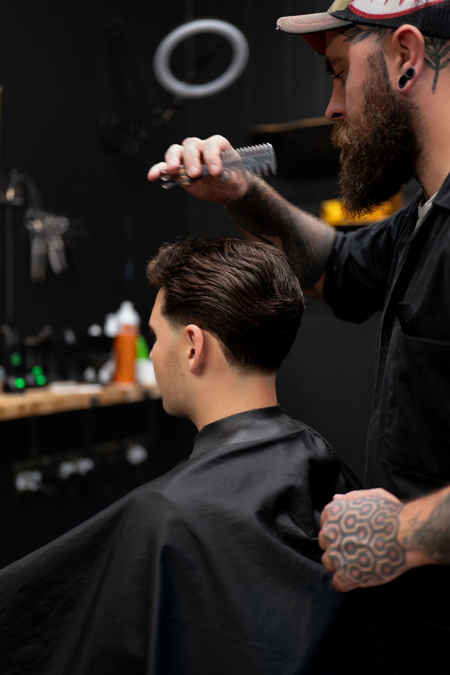 Tattooed bearded barber combing a young client's hair in a dark modern barbershop.