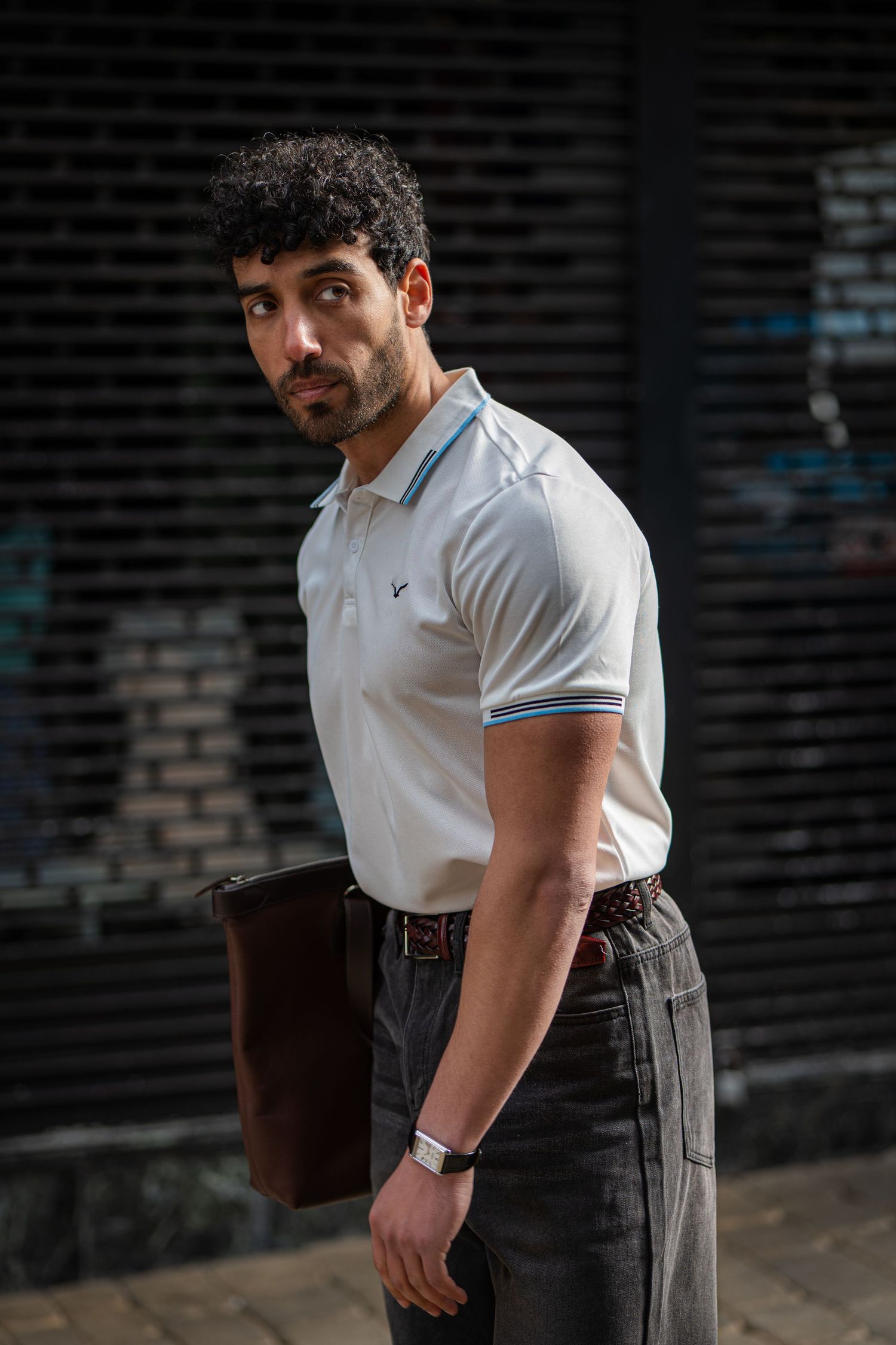 Athletic man in white polo shirt holding brown leather folder standing against dark urban background.