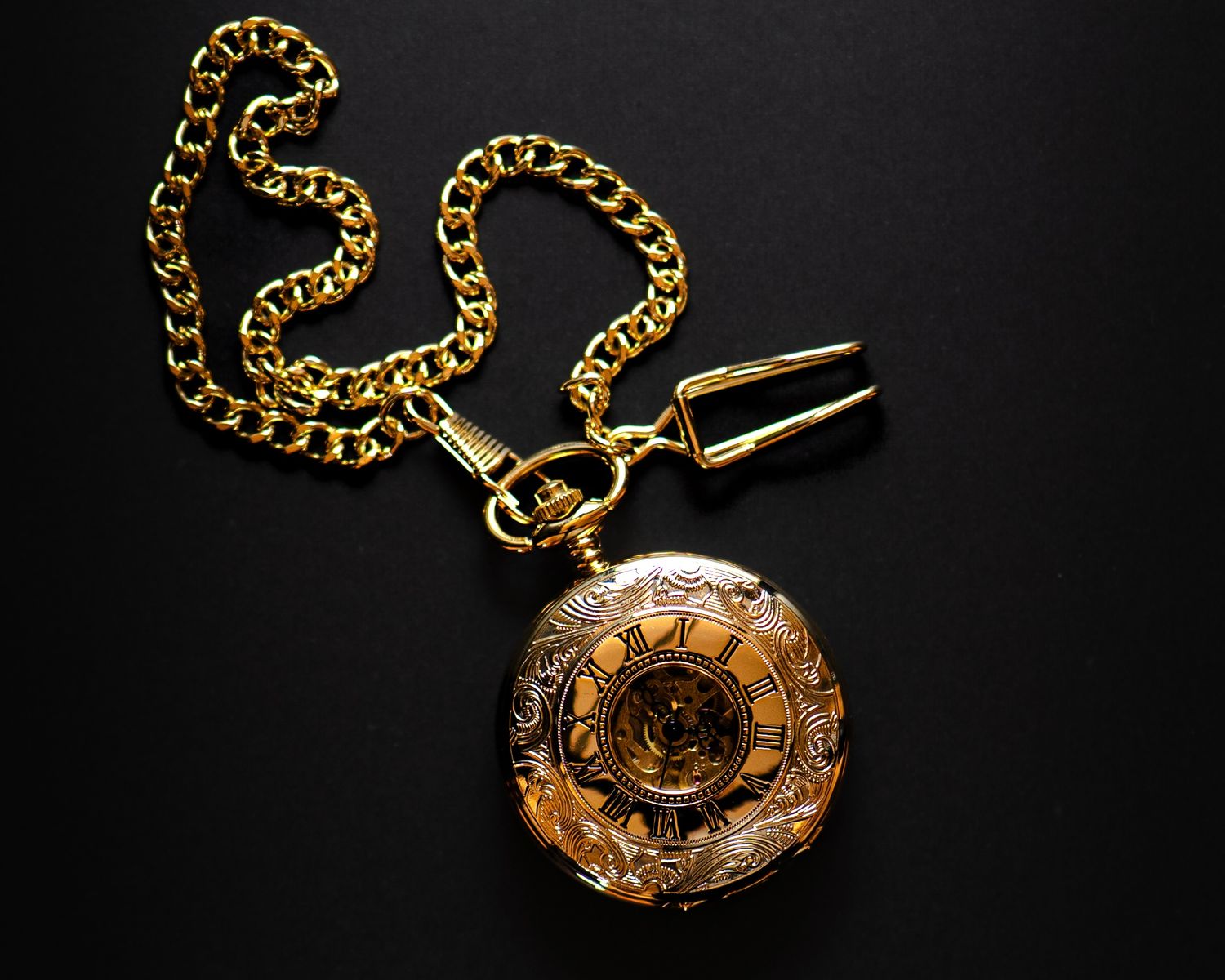 Ornate gold pocket watch with Roman numerals on a gold chain against a black background.