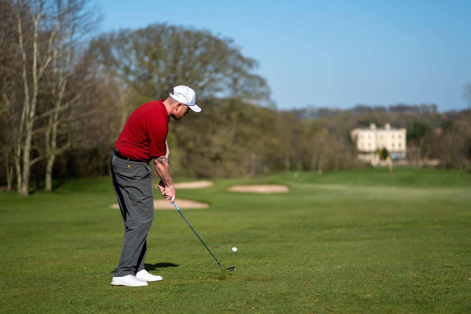 Male golfer in red sweater swinging a club on a scenic golf course with trees.