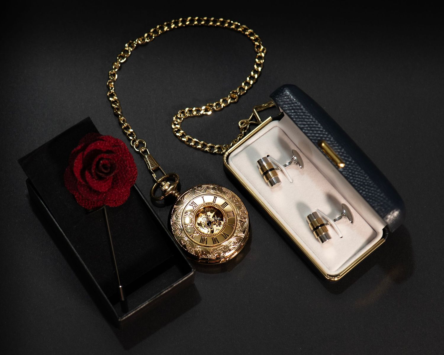 Gold pocket watch with chain beside red rose, black box, and cufflinks set on dark surface.
