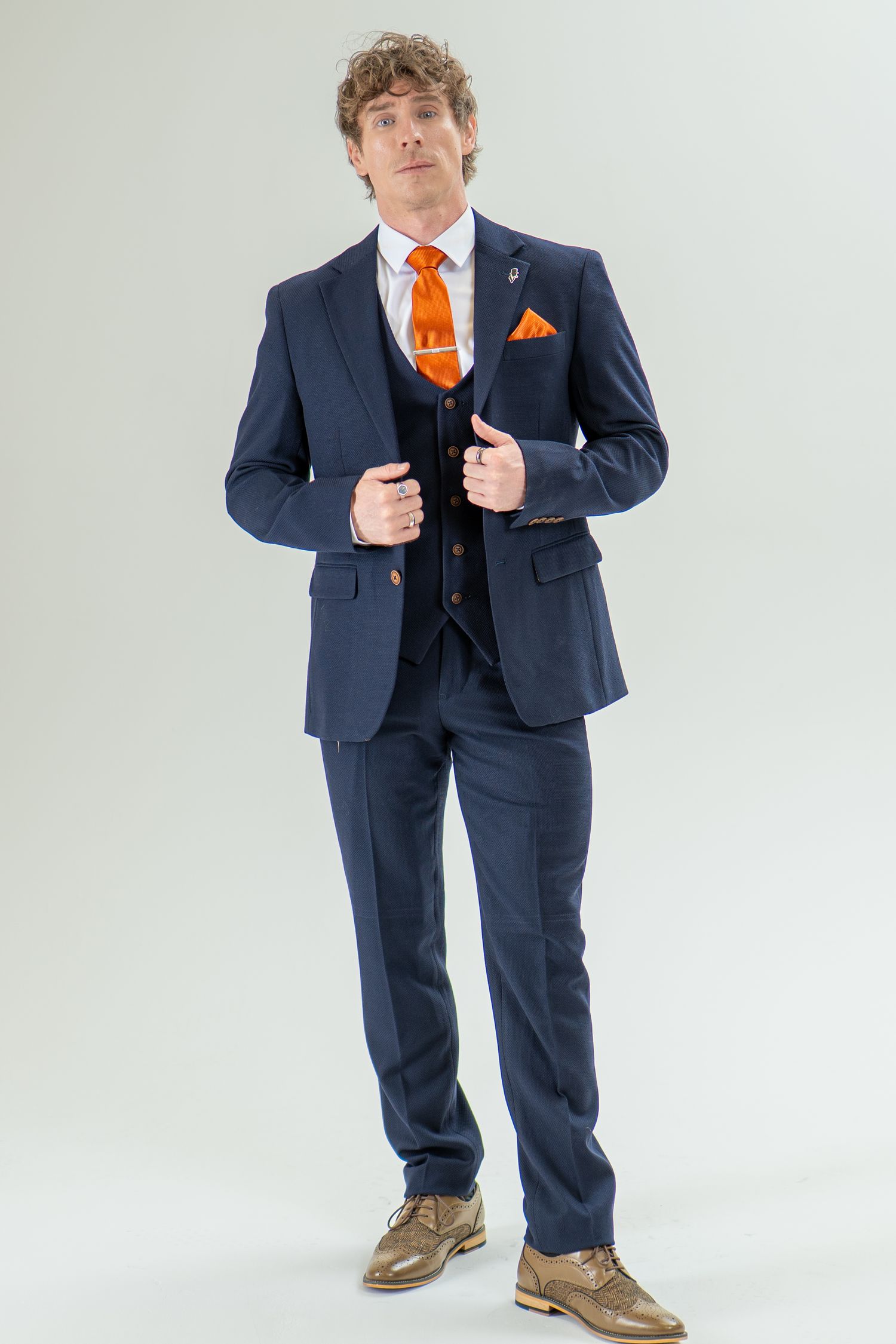 Man in navy blue three-piece suit with orange tie adjusting lapels, wearing tan brogues.