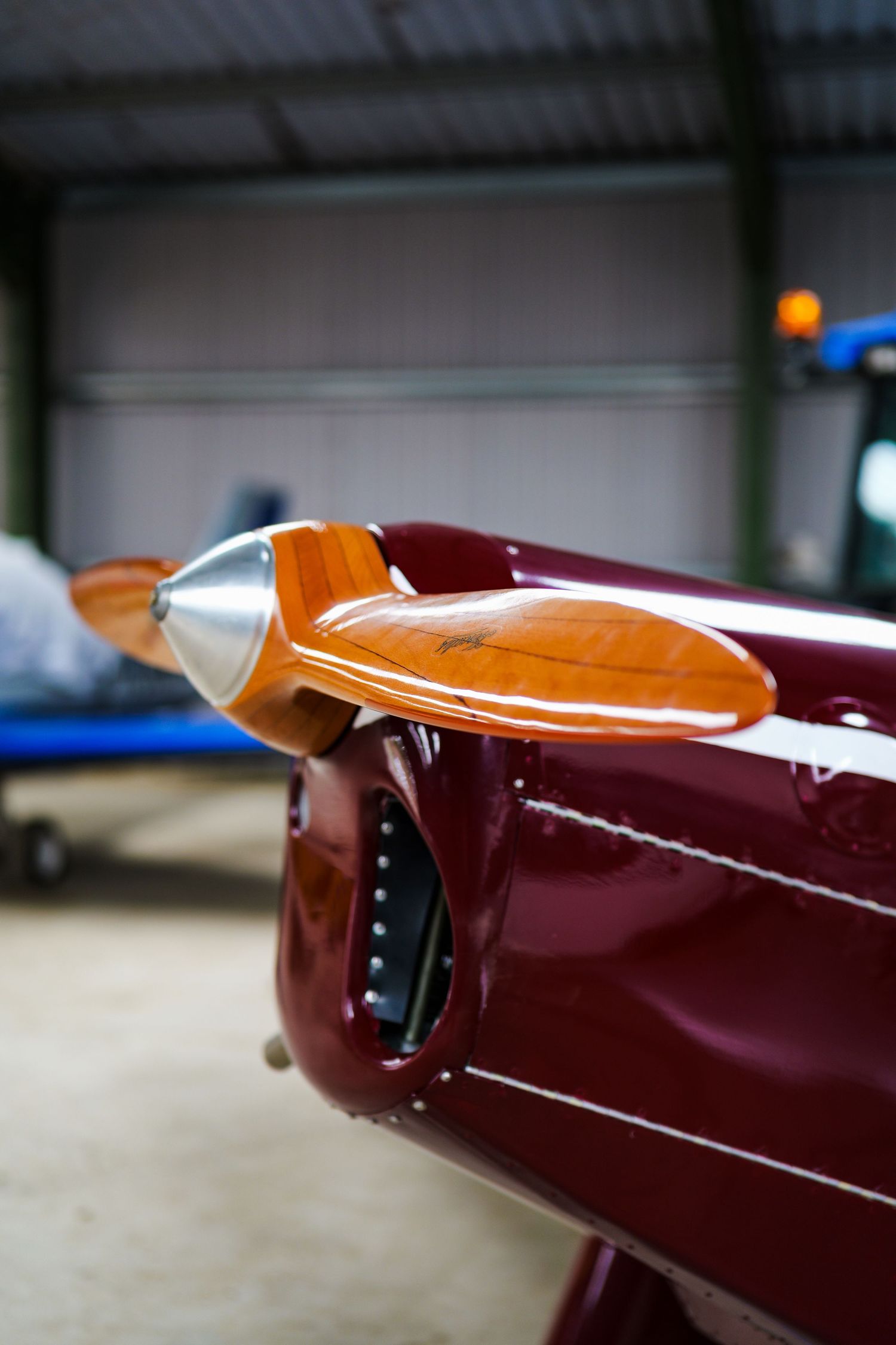Close-up of a polished dark red aircraft nose with a wooden propeller inside an aviation hangar.