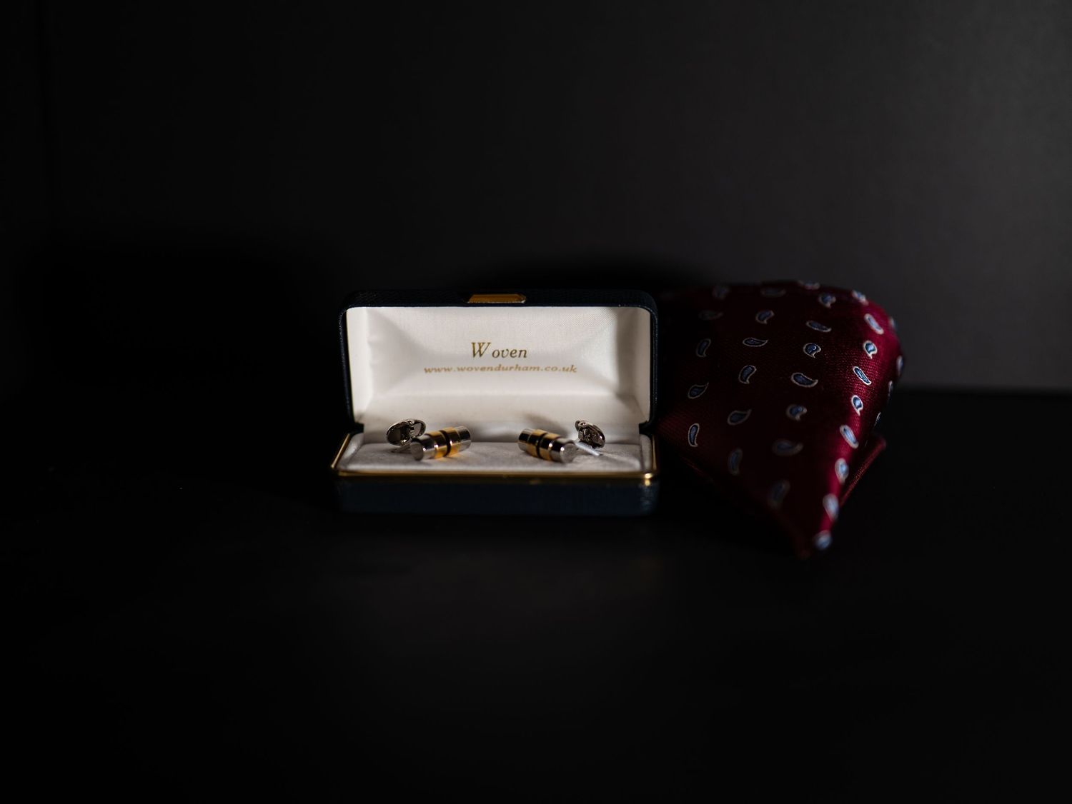 Gold and tortoiseshell cufflinks in white Weave jewelry box beside folded burgundy patterned tie.
