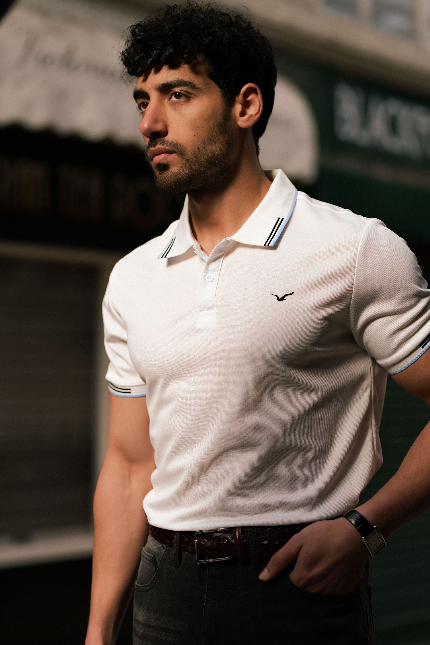 Stylish man wearing white polo shirt with navy tipped collar, dark jeans and brown belt posing in urban setting.