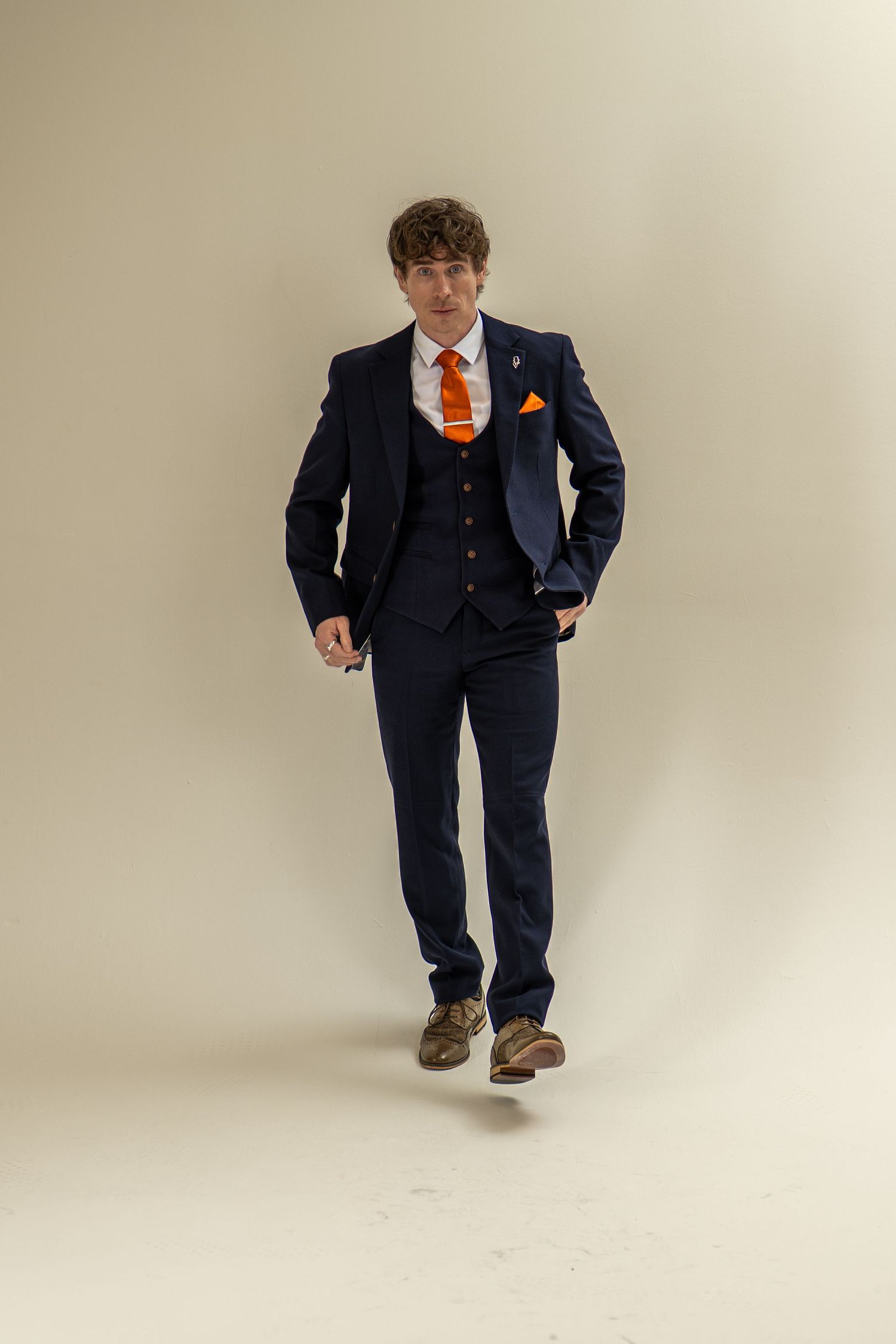 Young man in navy three-piece suit with orange tie and pocket square walking toward camera.