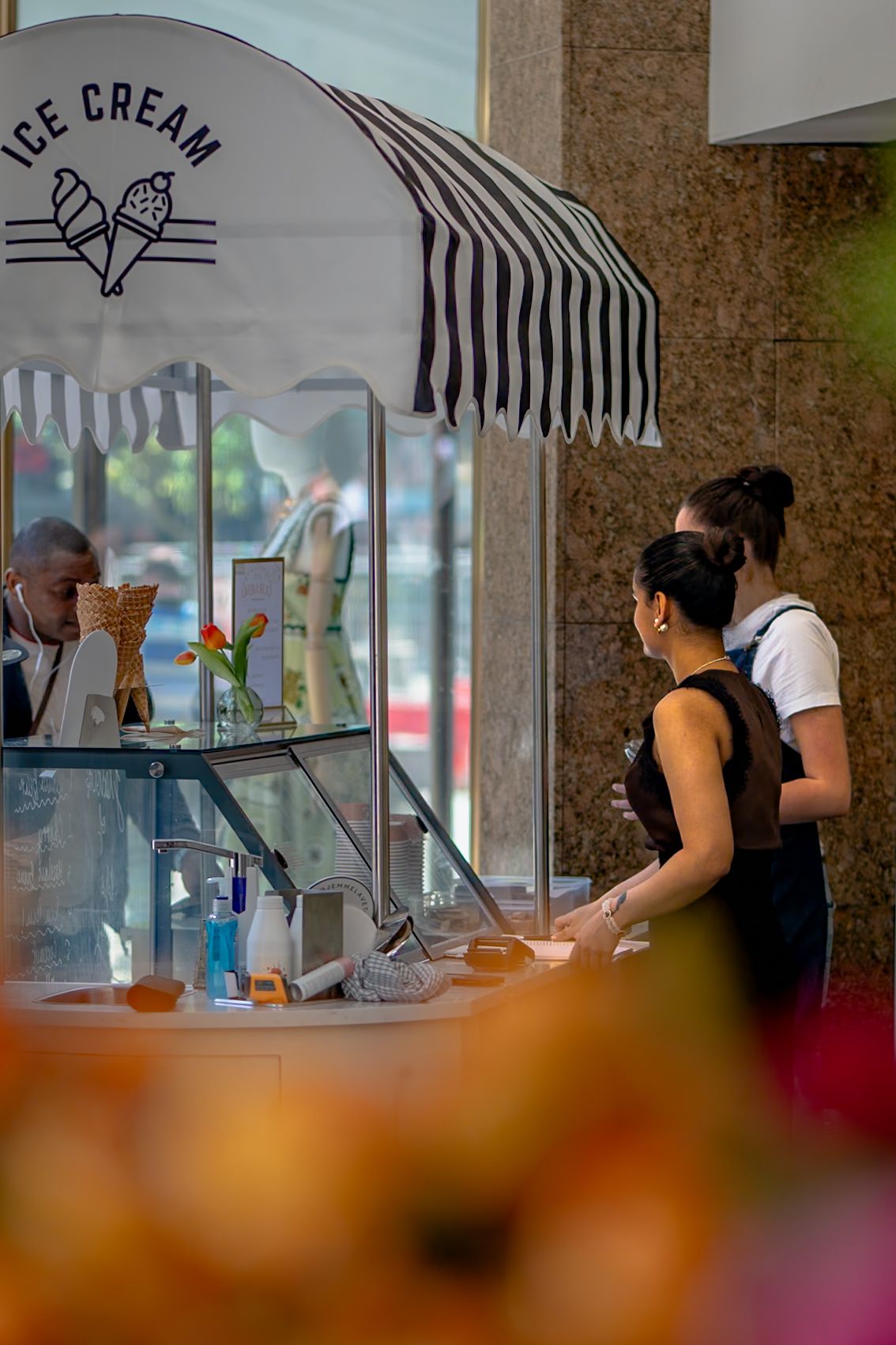 Two women stand at an outdoor ice cream cart with a striped awning while a vendor assists them.