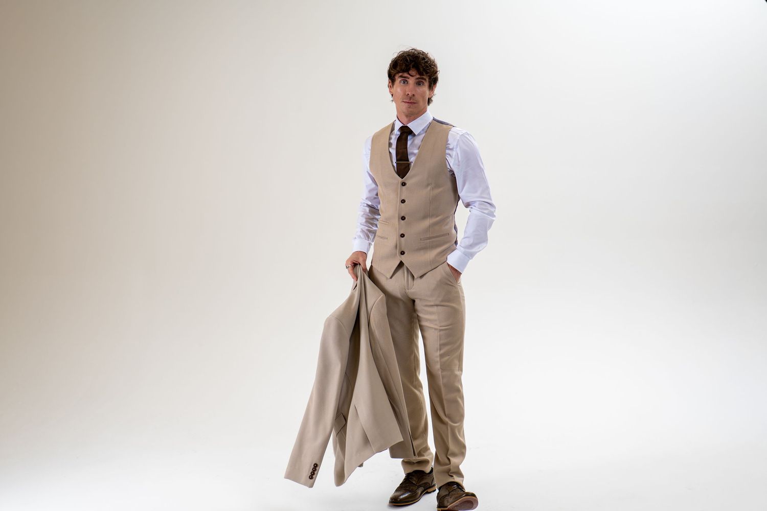 Stylish young man in beige three-piece suit holding jacket casually against neutral background.