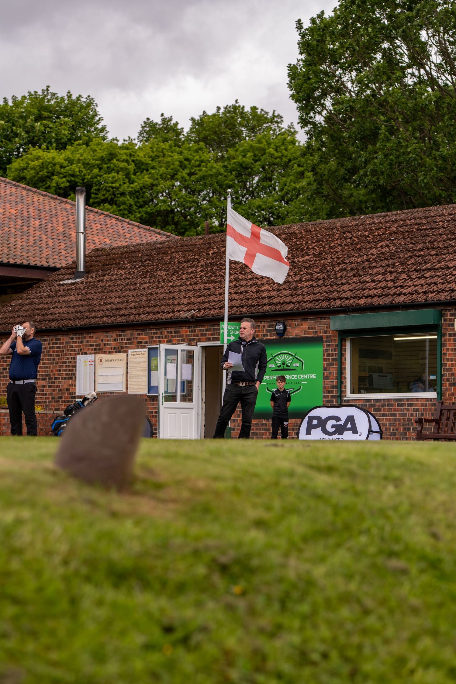 Golfer holding flag near PGA branded brick clubhouse with English St Georges Cross flag flying.