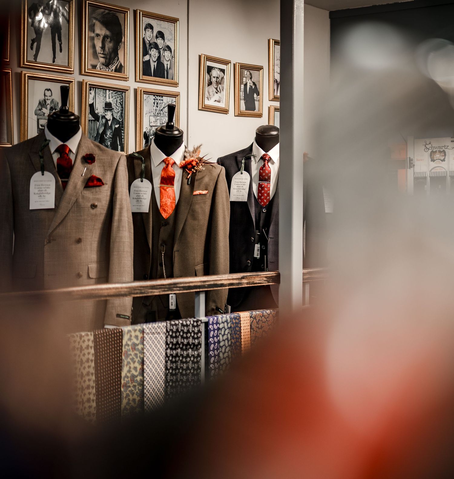 Three mannequins display tailored suits with red and orange ties in a menswear boutique with framed photos on the wall.