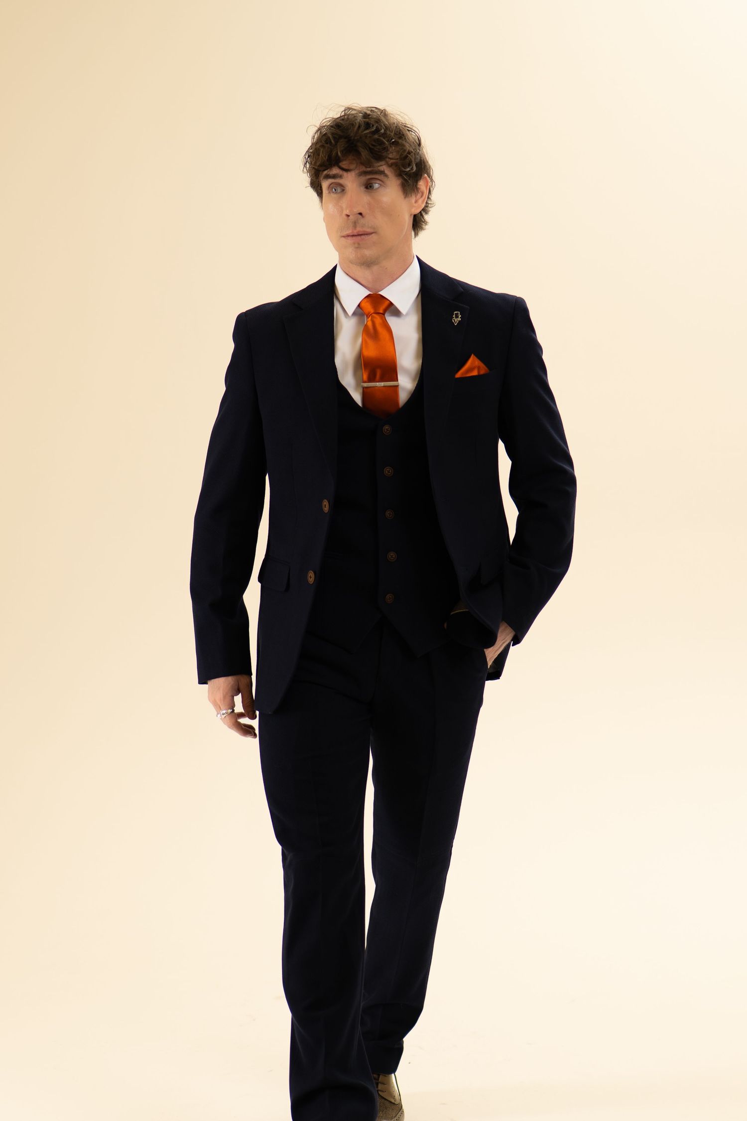 Tall man in dark navy three-piece suit with bold orange tie and matching pocket square, walking.