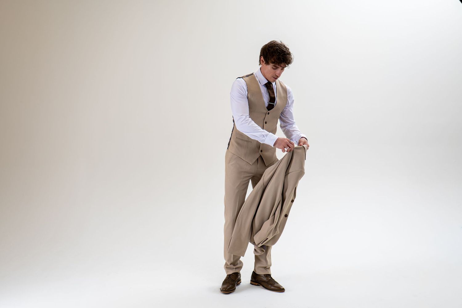 Young man in beige waistcoat and trousers holding suit jacket while looking down.