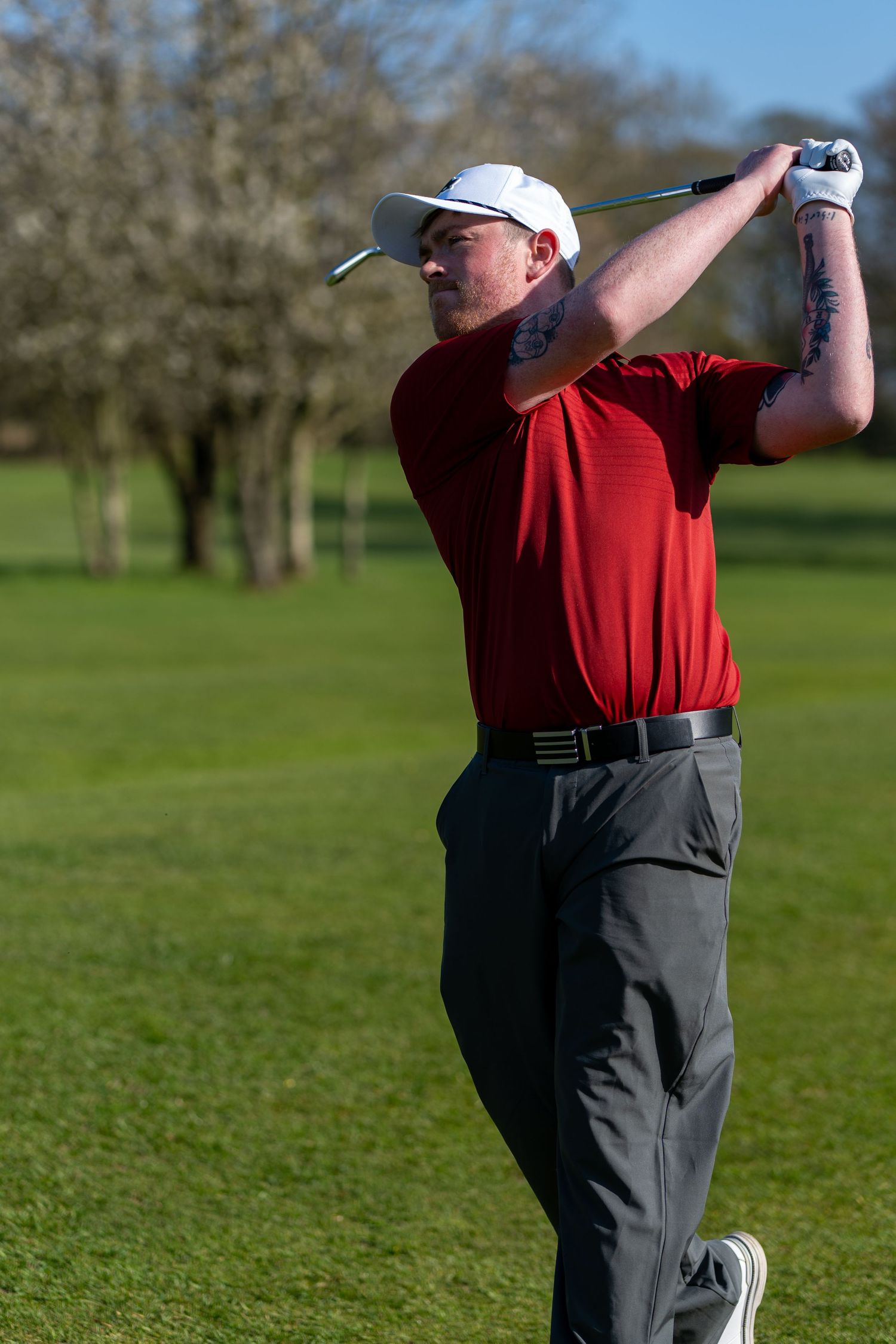 Male golfer in red shirt and white cap completing follow-through swing on sunny golf course with trees behind.