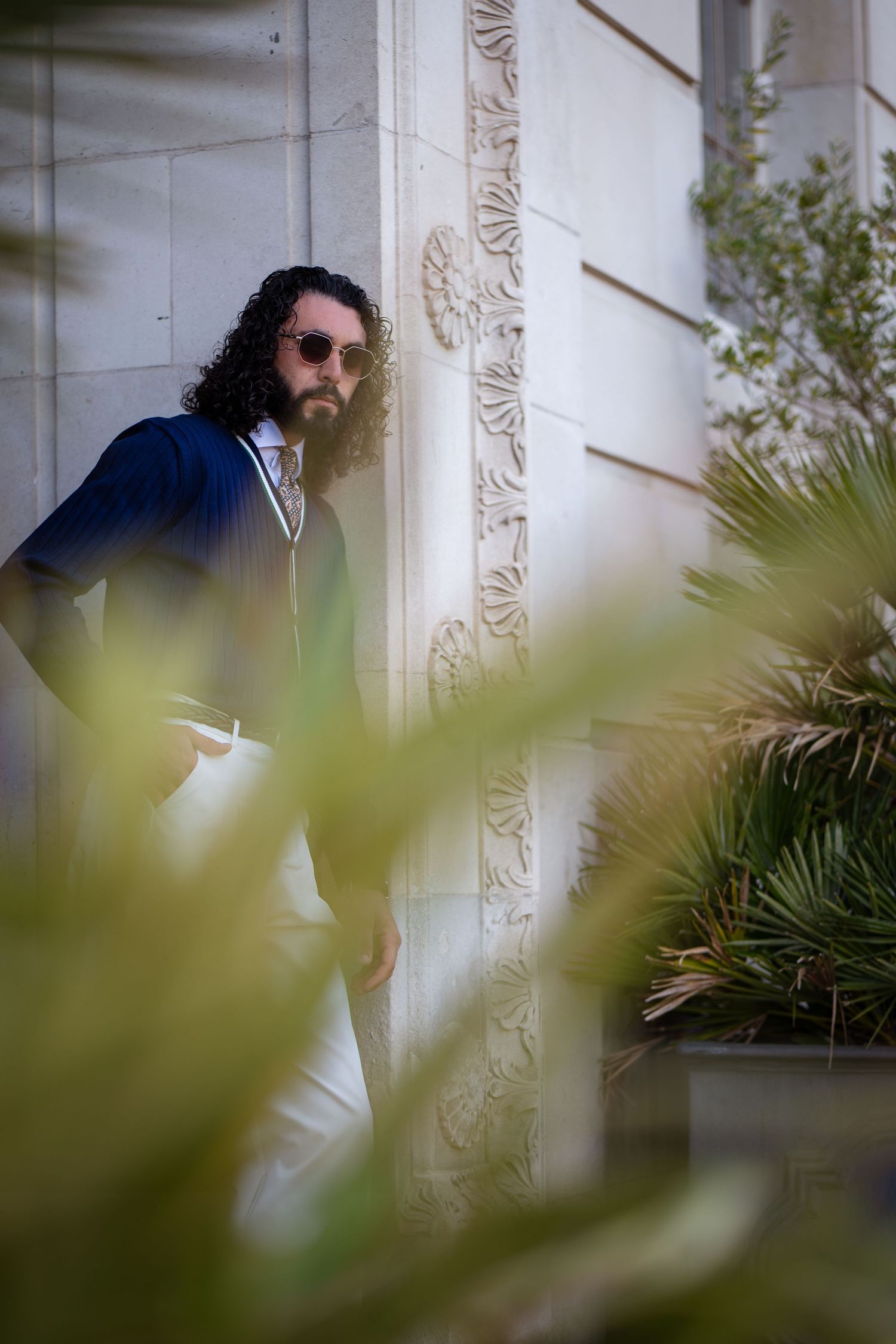 Man with curly hair and sunglasses in navy cardigan and white trousers leaning against ornate building wall.