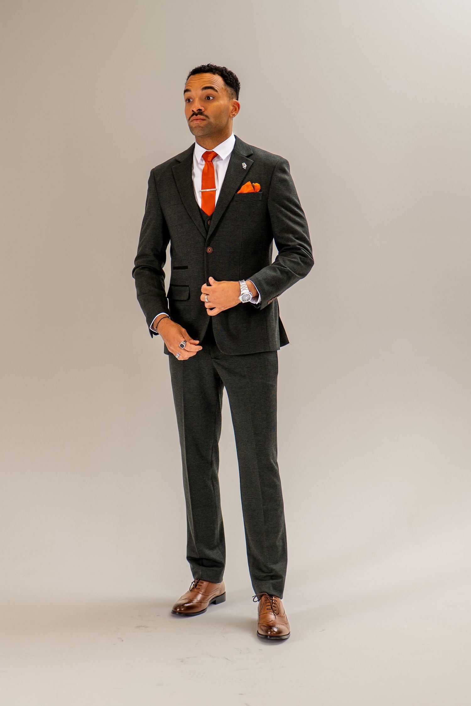 Confident man in a charcoal three-piece suit with orange tie and brown leather shoes posing.