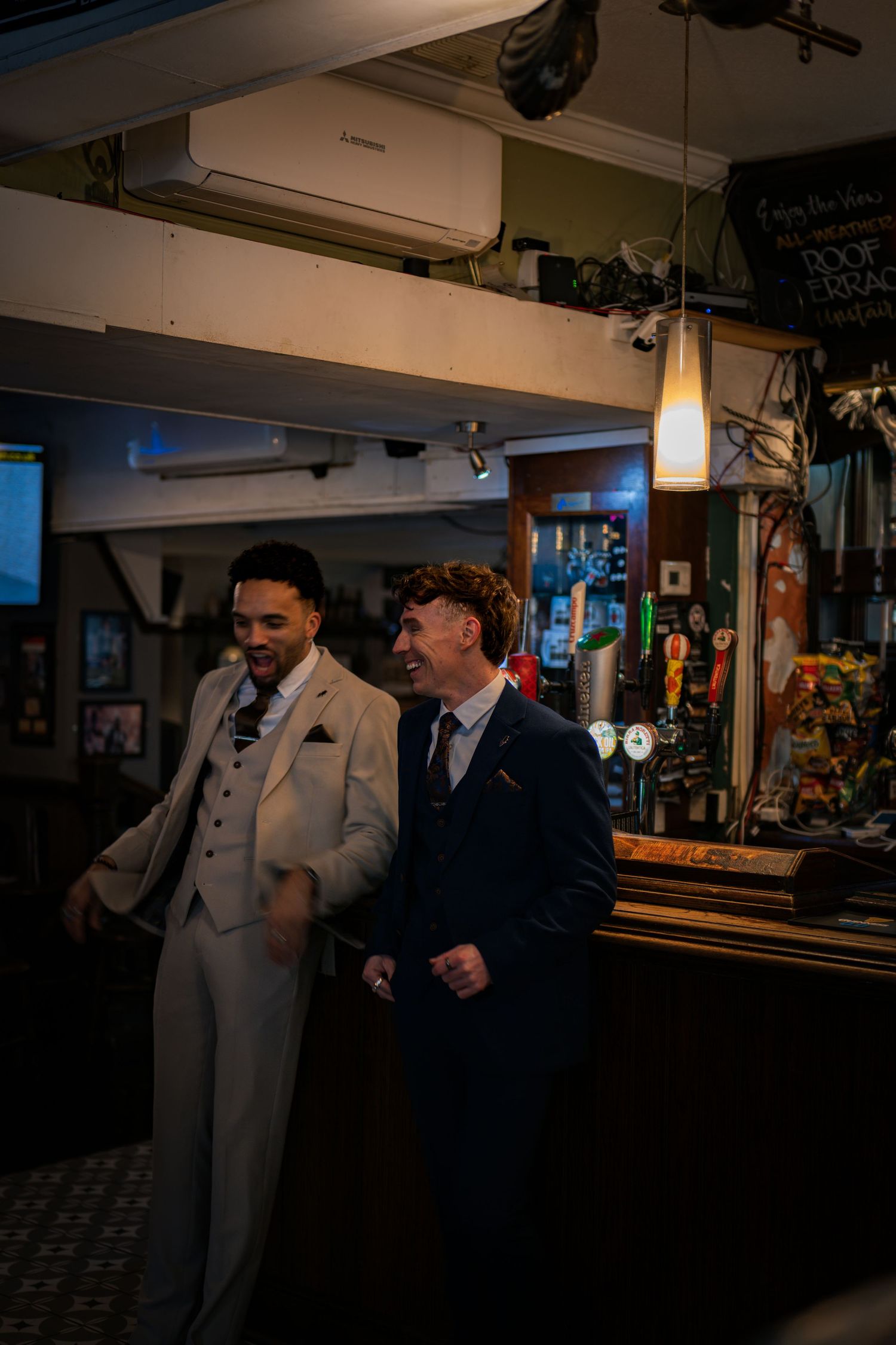 Two smartly dressed men in suits laughing together at a pub bar during a celebration.