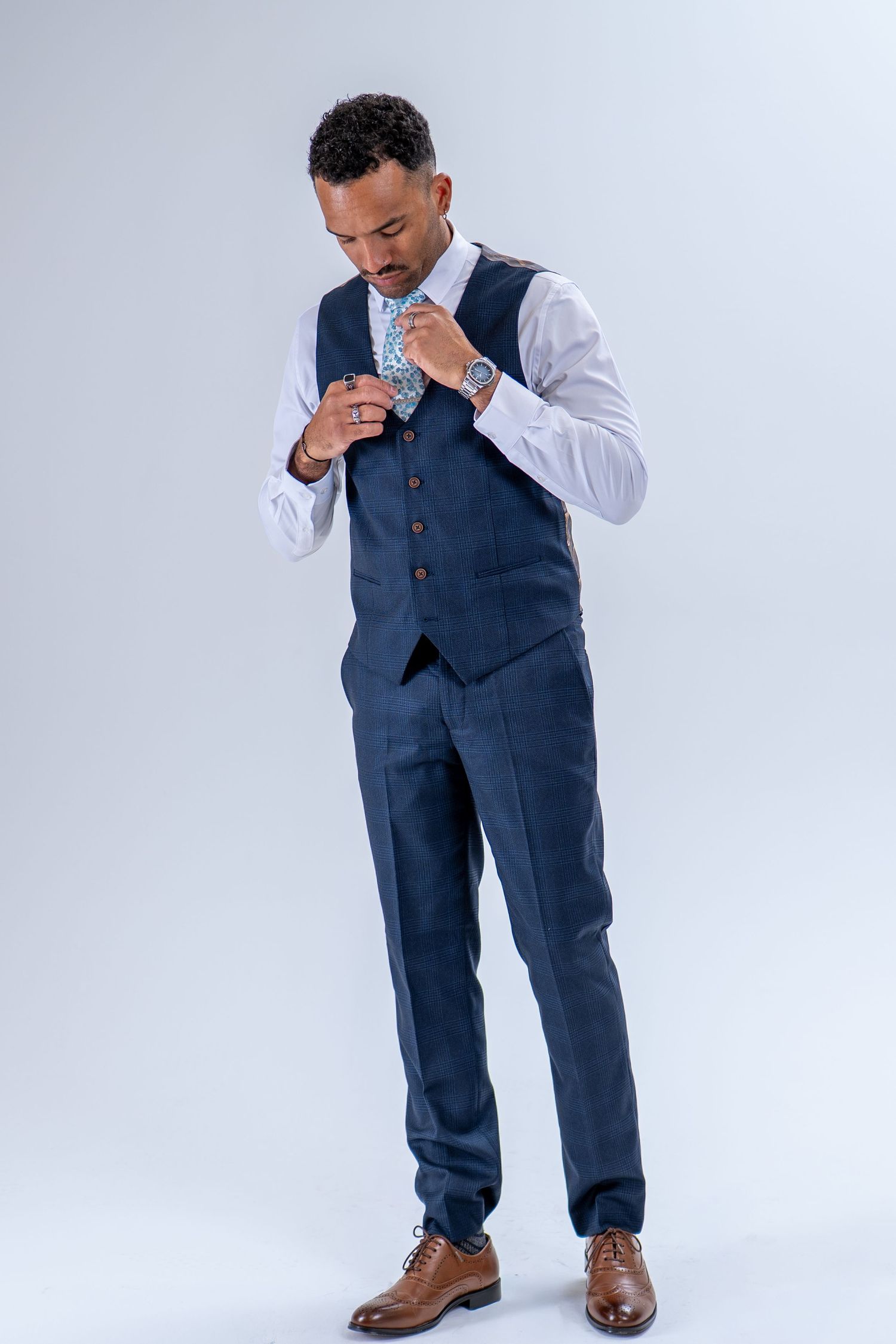 Well-dressed man in navy blue waistcoat adjusting floral tie with luxury watch on wrist.
