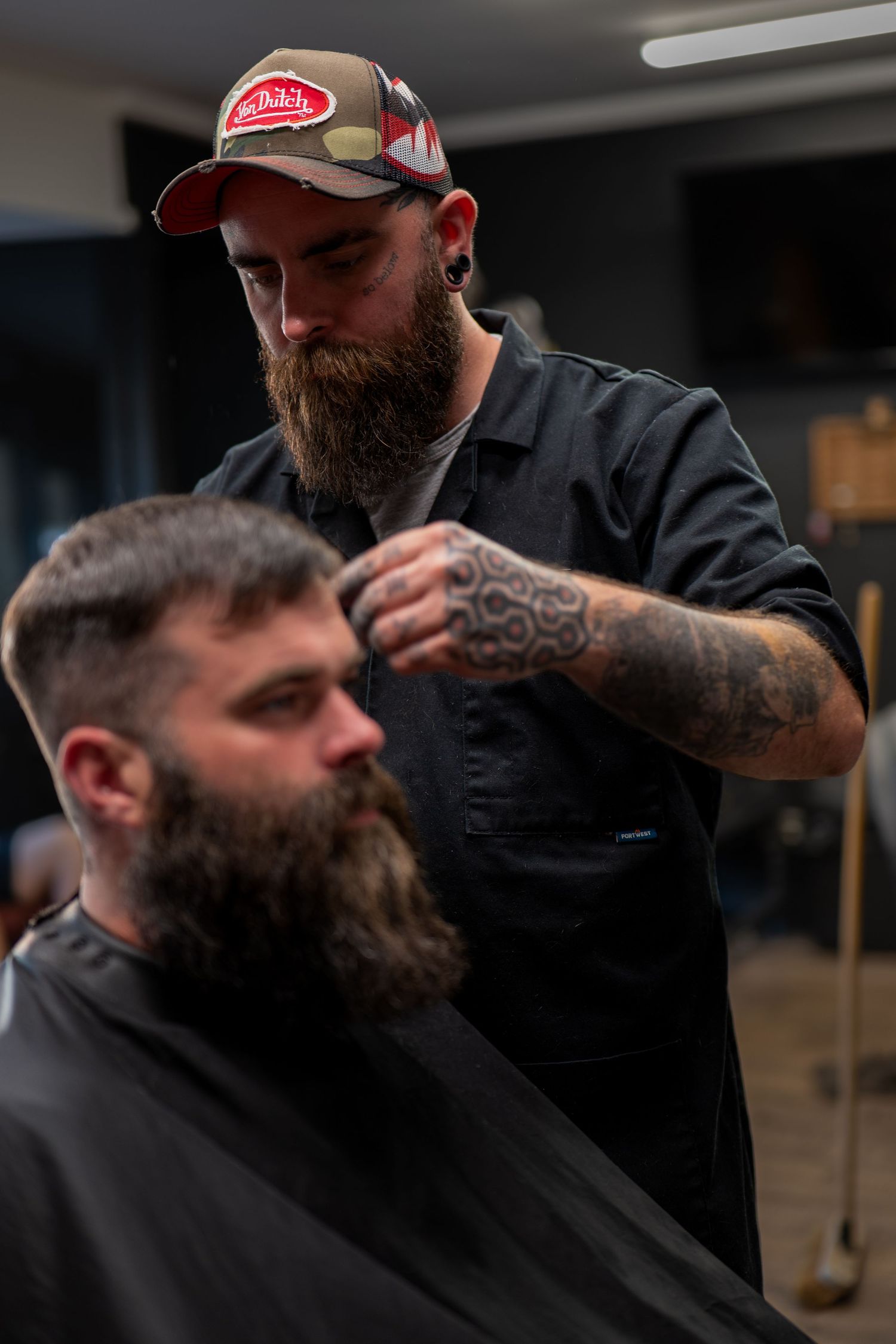 Tattooed barber with beard and cap cutting a bearded client's hair in a barbershop.
