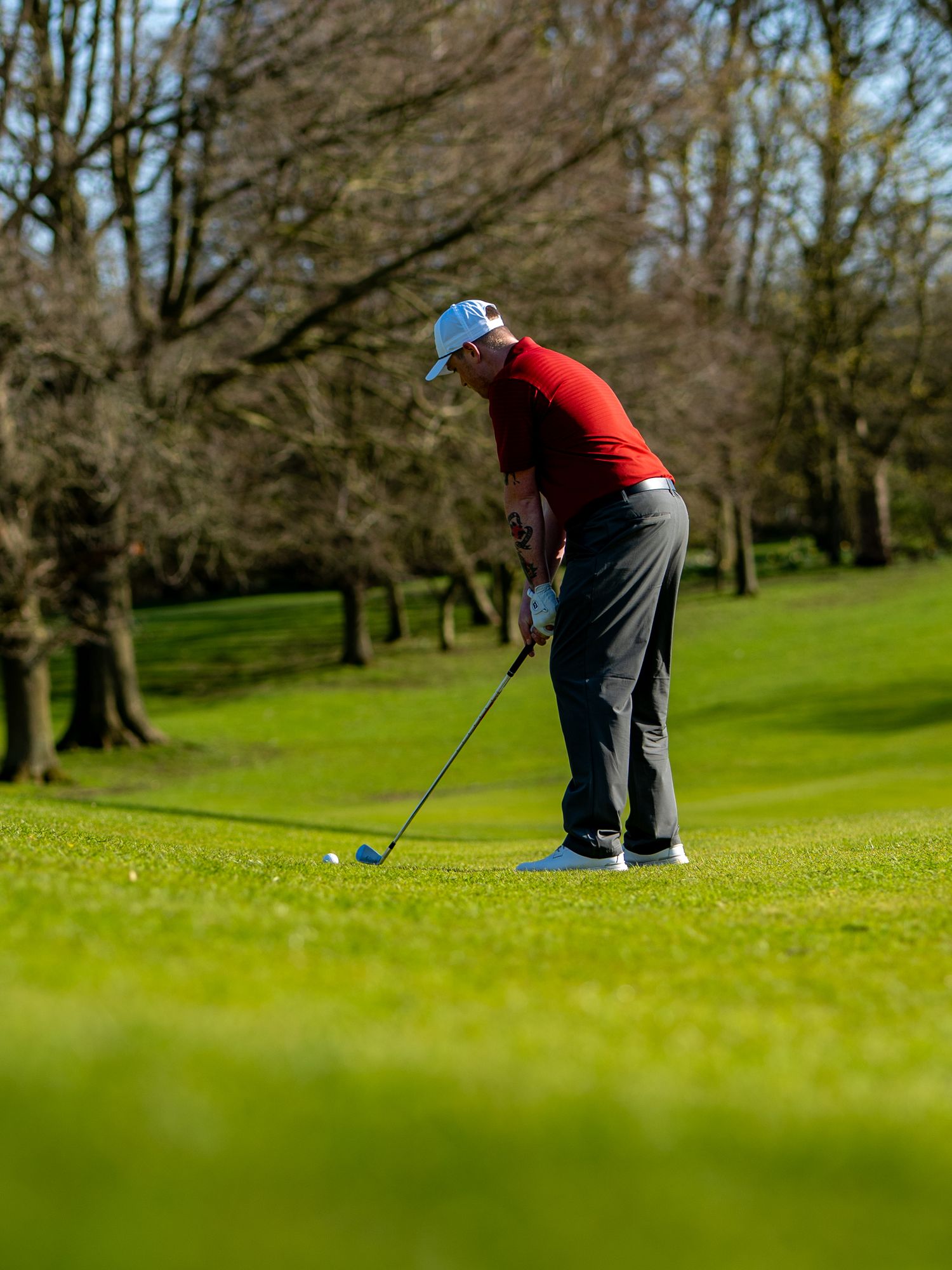 Golfer in red shirt and blue cap preparing to strike ball on lush green golf course surrounded by trees.